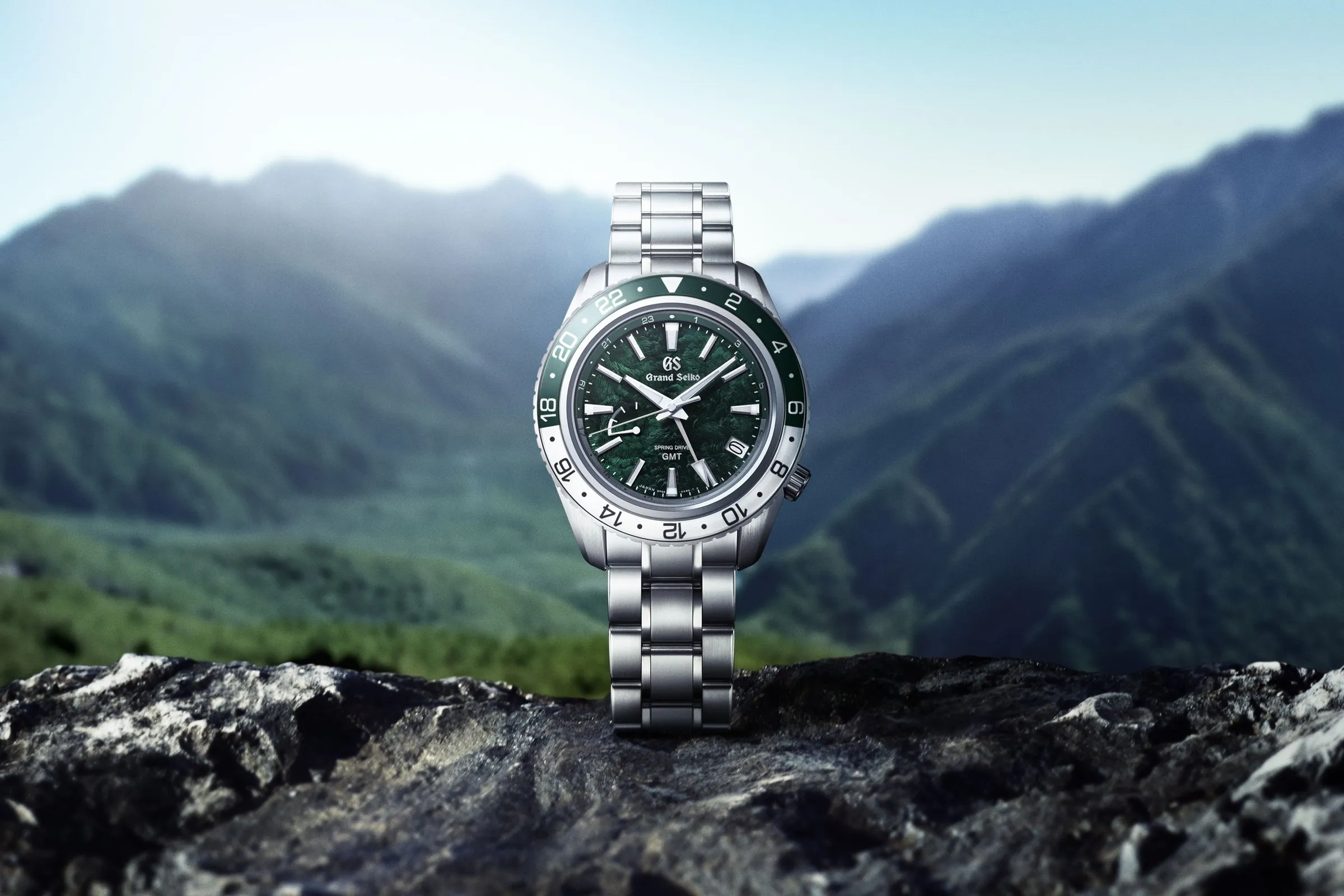 A Grand Seiko Spring Drive GMT watch with a green dial, a green and white bezel and a steel bracelet, standing on a dark rock with misty green mountains behind it