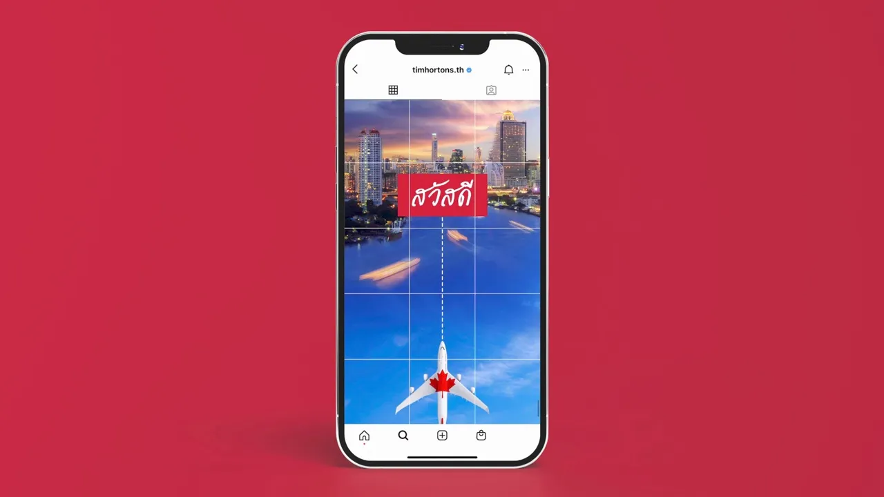 หน้าจอโทรศัพท์แสดงหน้า Instagram ของ Tim Hortons Thailand เครื่องบินที่มีใบเมเปิลสีแดงบินจากภูเขาในแคนาดามาถึงตึกสูงริมแม่น้ำในกรุงเทพฯ พร้อมคำว่า Hello ที่เปลี่ยนเป็นคำว่าสวัสดี