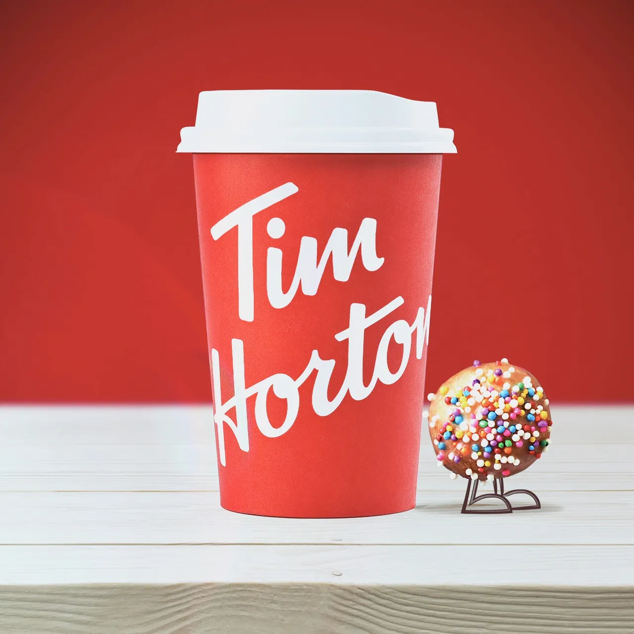 แก้วสีแดงของ Tim Hortons บนโต๊ะสีขาว มี Timbit โรยเม็ดน้ำตาลหลากสีที่วาดแขนขาเดินออกมาจากหลังแก้วและโบกมือ ใต้คำว่าสวัสดี