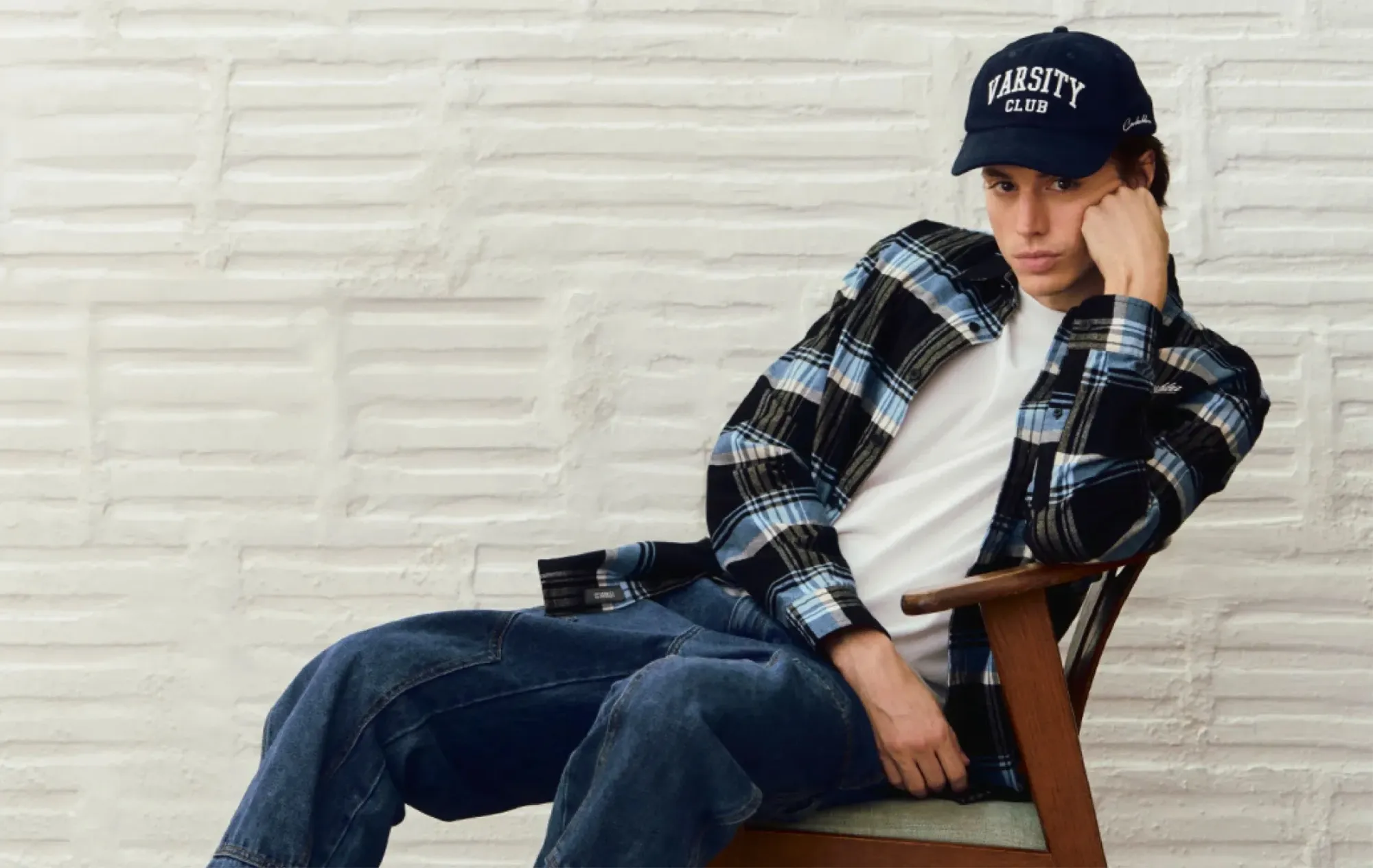 A young man in a navy cap, a blue and black check shirt over a white T-shirt and wide blue jeans, leaning back in a wooden armchair against a white plaster wall
