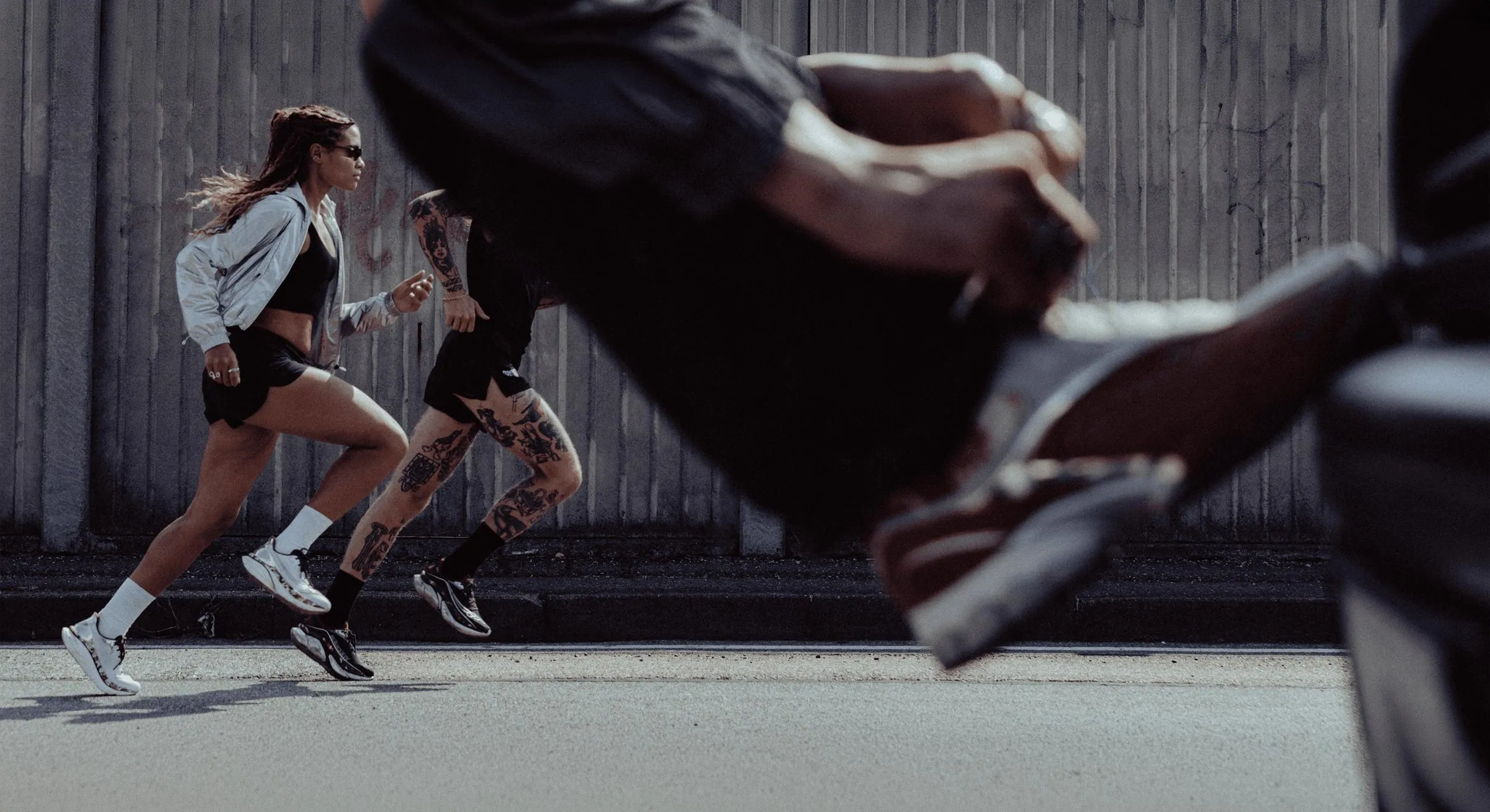 2 runners in black shorts running along a road in front of a grey wall, one in a silver jacket and sunglasses and the other with tattooed legs, with the blurred arm and shoe of another runner in the foreground