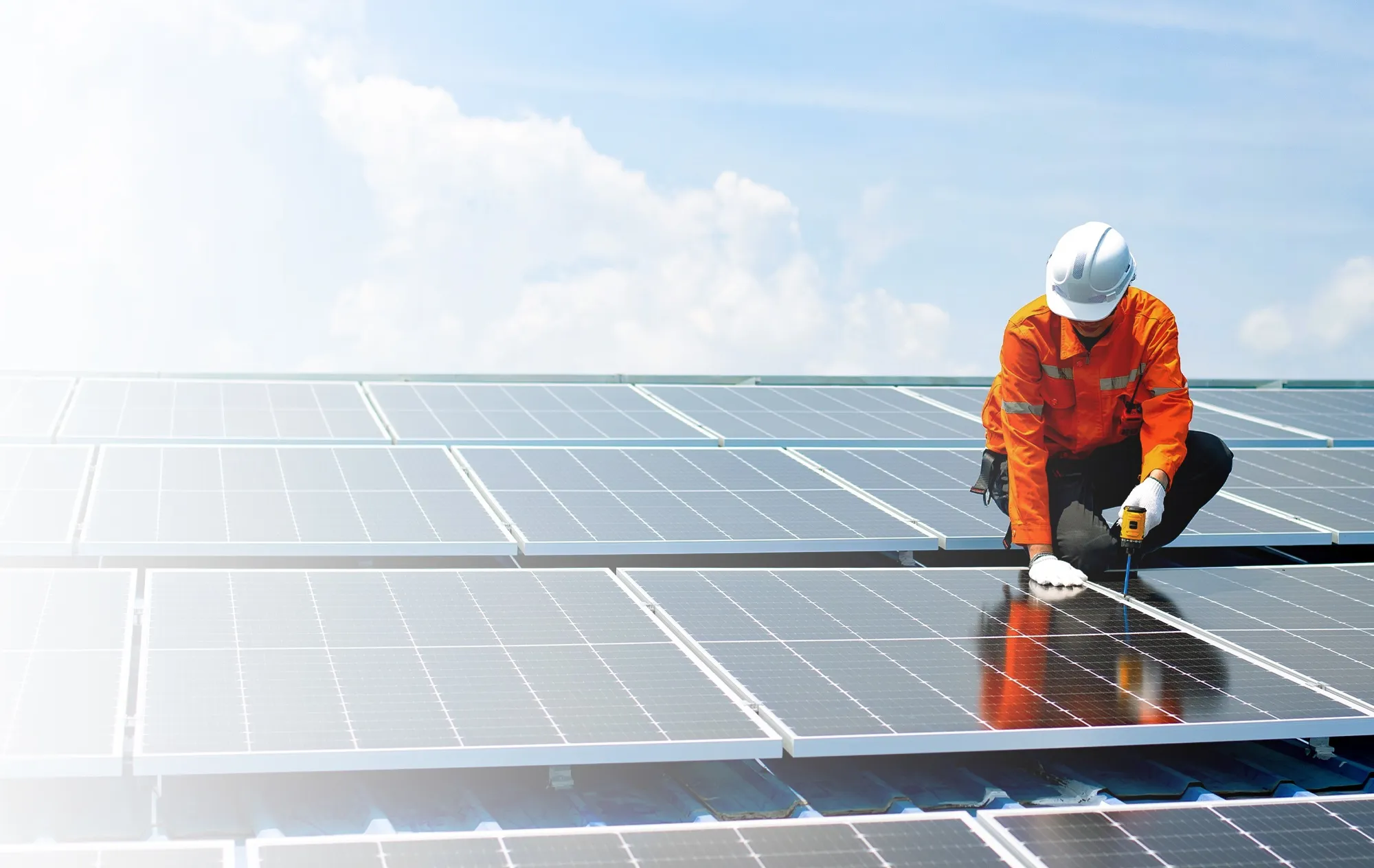 A worker in an orange uniform and a white hard hat fixing a solar panel with a power drill on a roof full of solar panels, under a blue sky