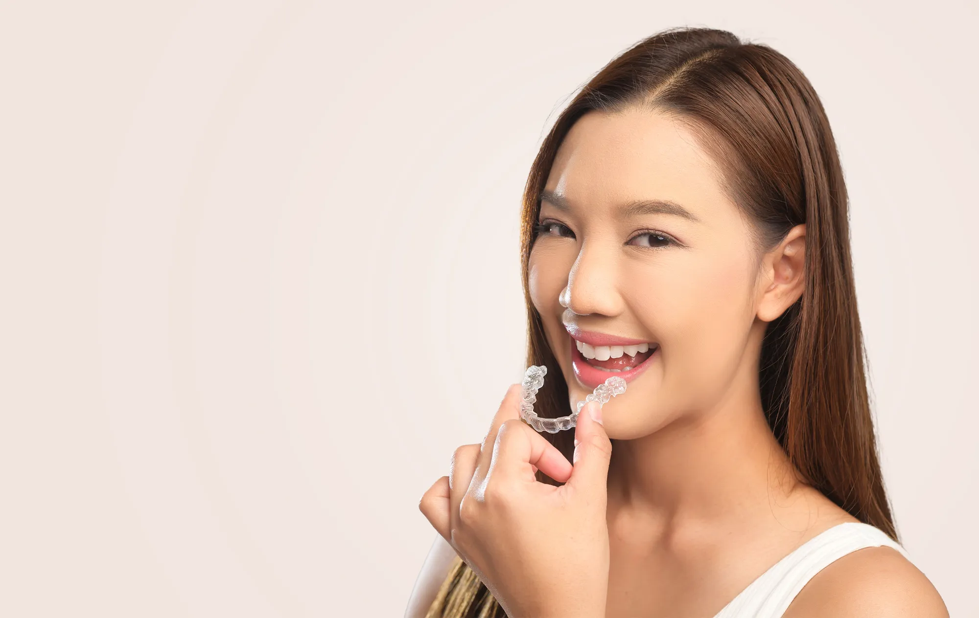 A smiling woman holding a clear aligner up to her teeth, against a pale pink background