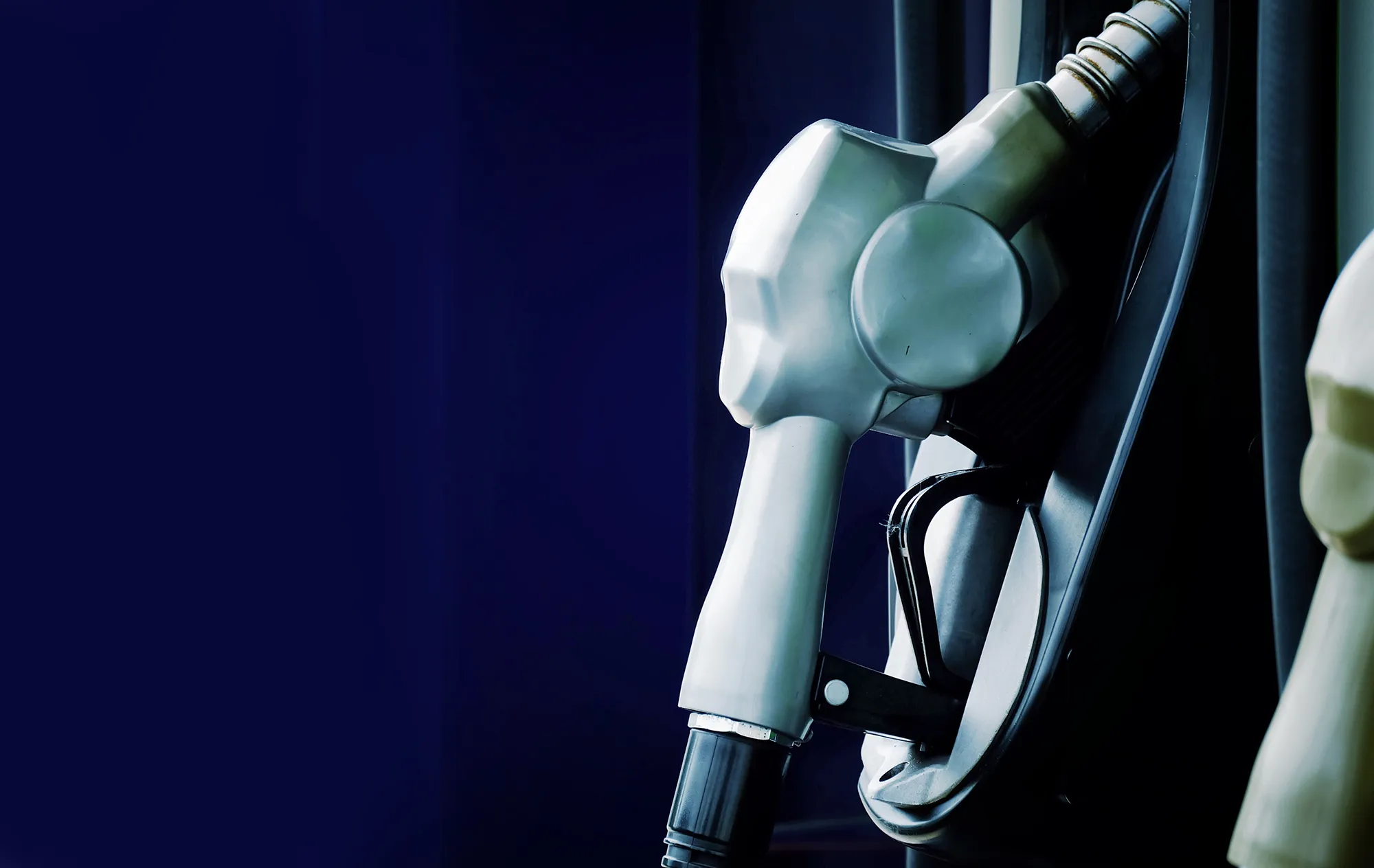 A silver fuel nozzle hanging in a pump holder against a dark blue background, the edge of a second nozzle beside it