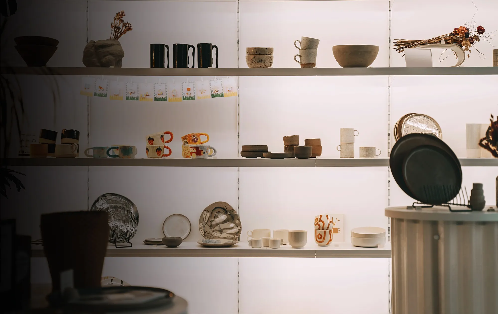 Backlit white shelves of ceramic mugs, bowls and plates, some hand-painted, with a row of small illustrated cards hanging on a string