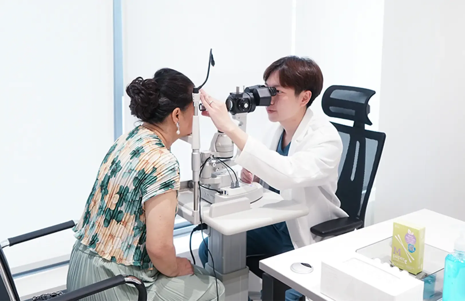 An ophthalmologist in a white coat looks through a slit lamp to examine the eyes of a woman in a floral top, in a bright white consultation room