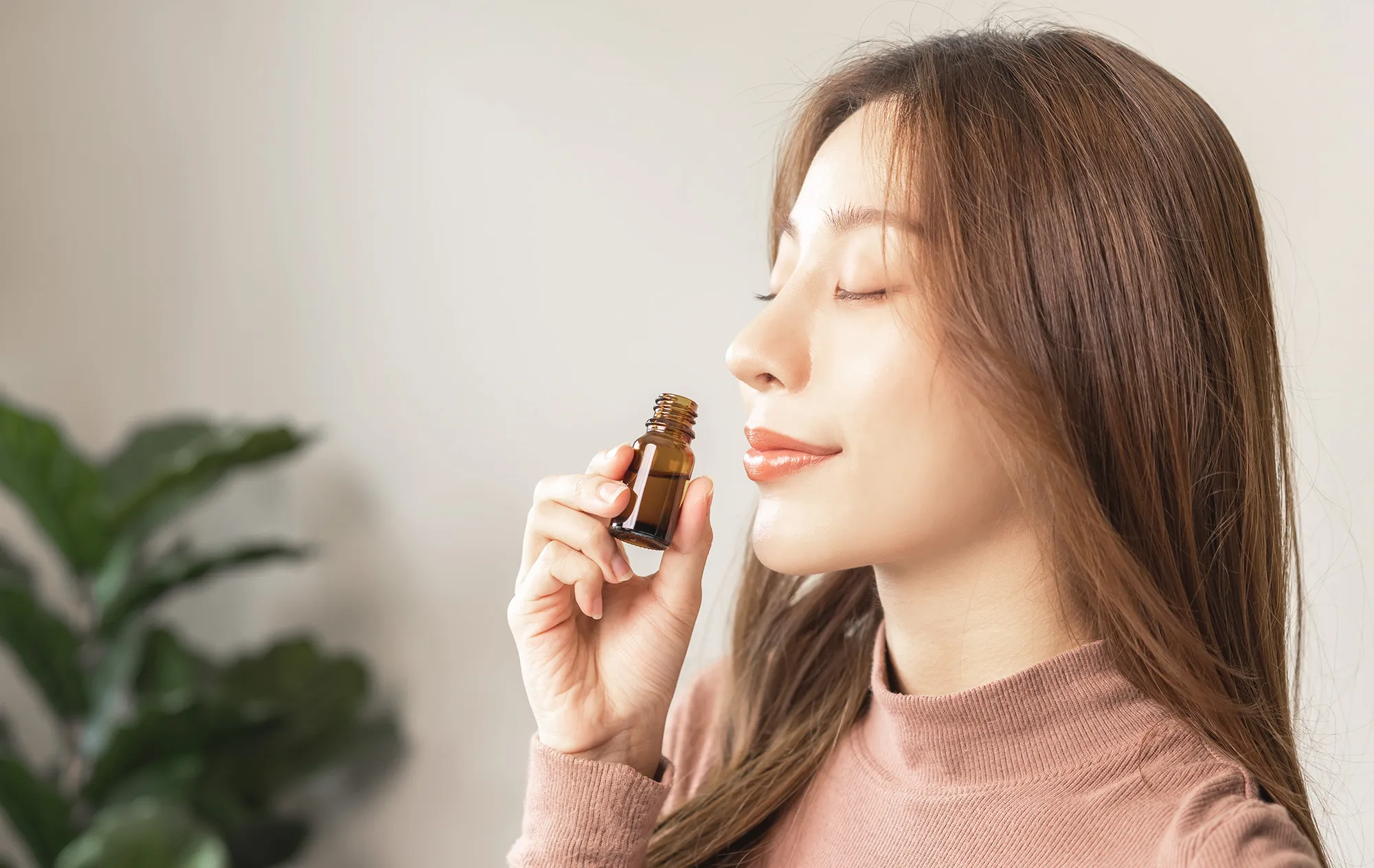 A woman with long brown hair and her eyes closed smelling a small open amber glass bottle, a green plant blurred to the left