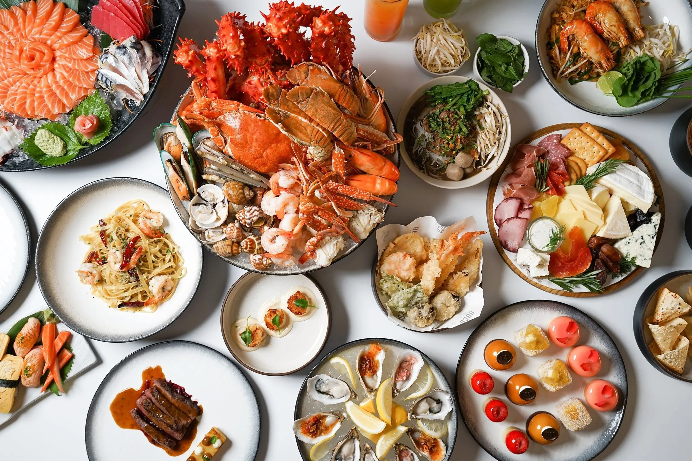 A white table seen from above, covered with dishes including a platter of crab, prawns and shellfish, salmon sashimi, pasta with prawns, tempura, a cheese and meat board, oysters and small desserts