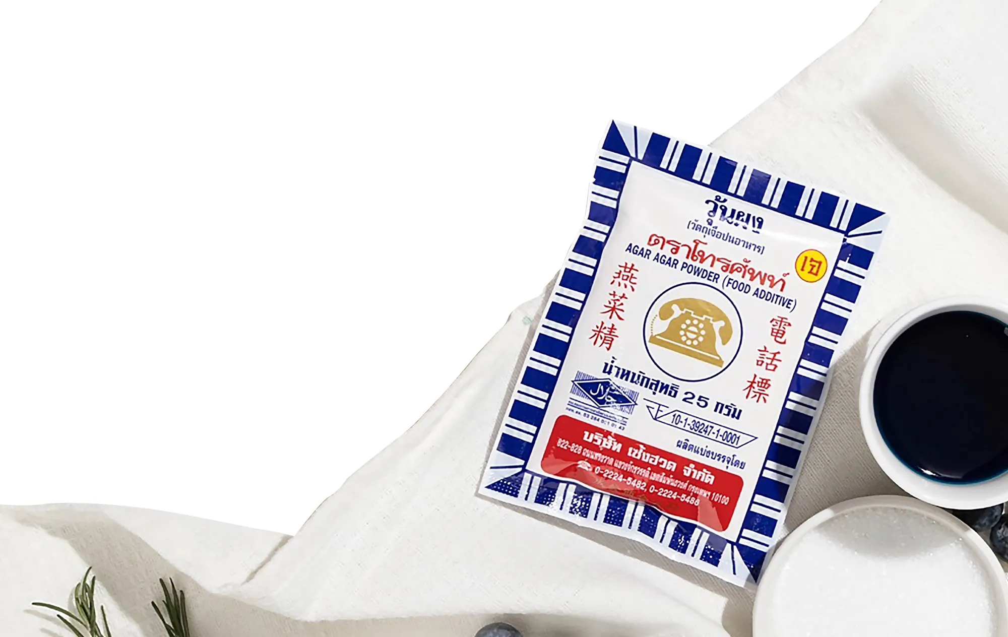A 25 gram sachet of Telephone brand agar powder on a white cloth, beside a cup of dark blue liquid, a bowl of white crystals and blueberries