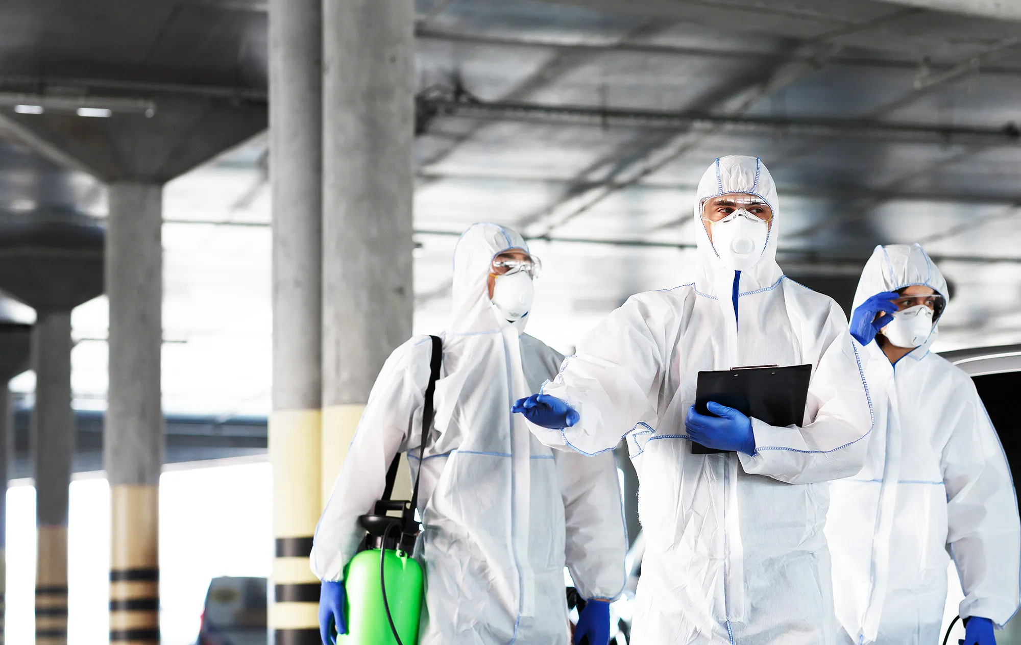 Three people in white protective suits, masks and goggles walking through a car park, one holding a clipboard and one carrying a green sprayer