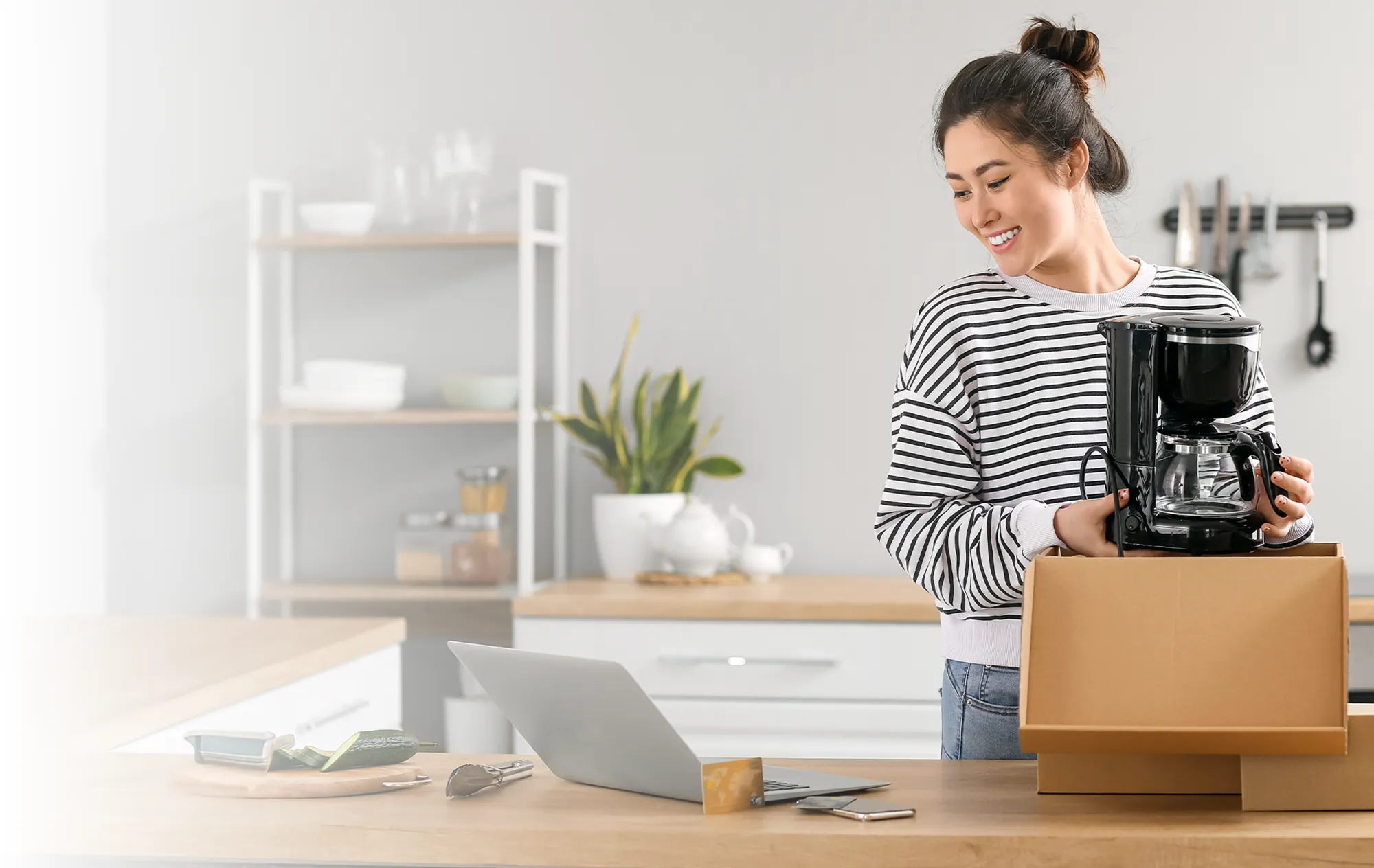 A woman in a striped top lifting a coffee maker out of a delivery box in a kitchen, a laptop open on the counter
