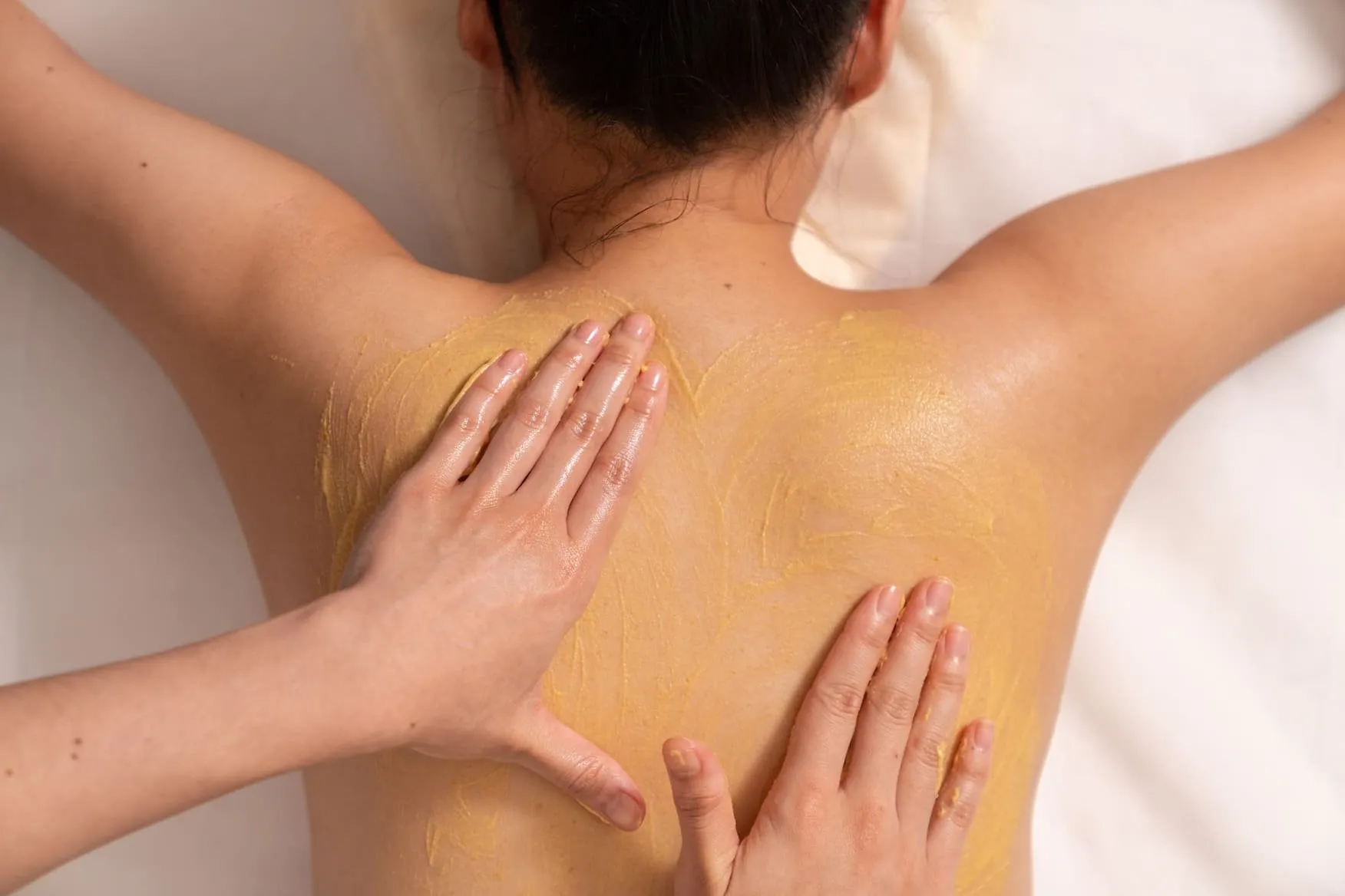 A woman lying face down seen from above, with a yellow scrub spread across her back and two hands massaging it