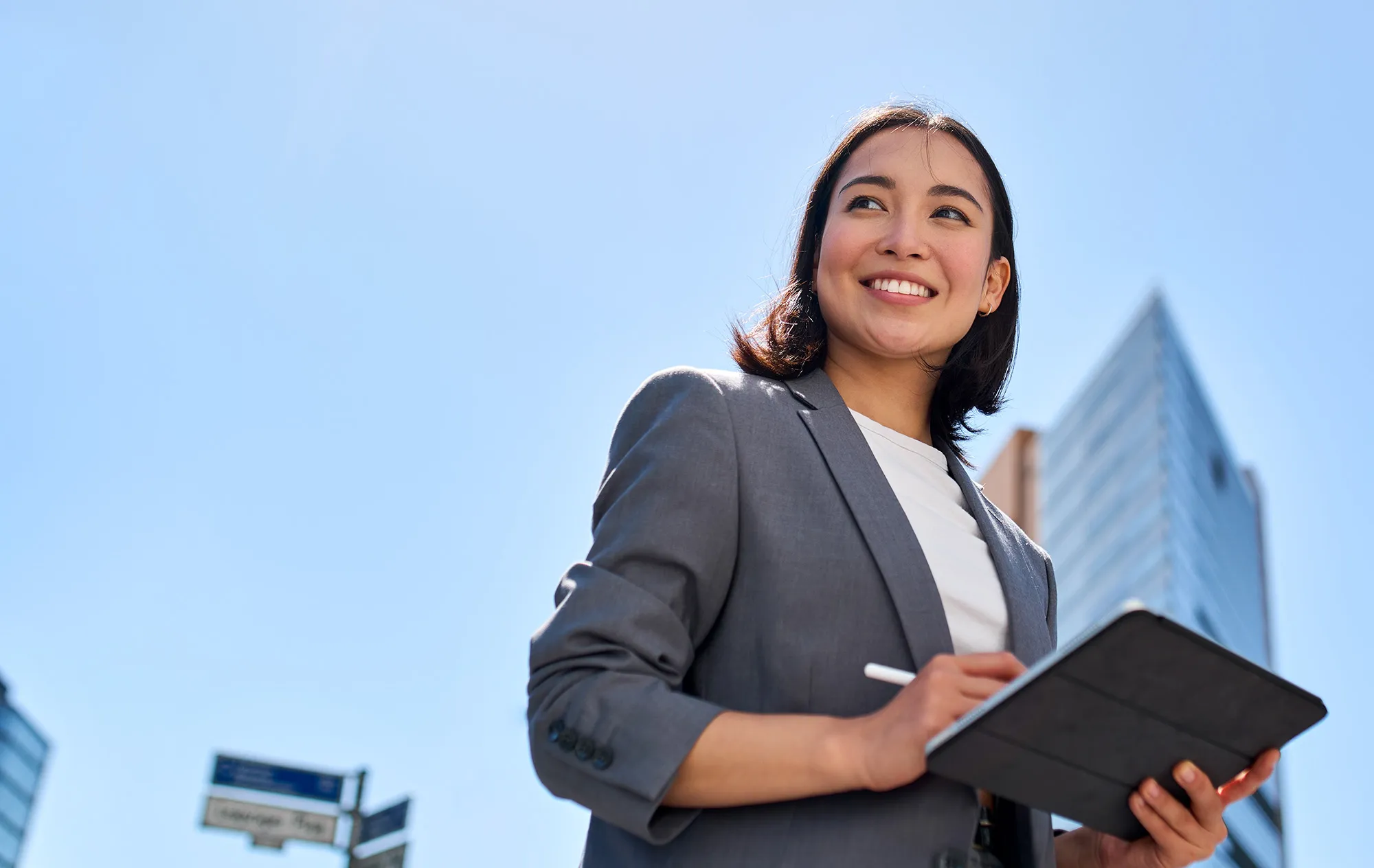A smiling woman in a grey blazer holding a tablet and a stylus outdoors, a glass office tower and a blue sky behind her