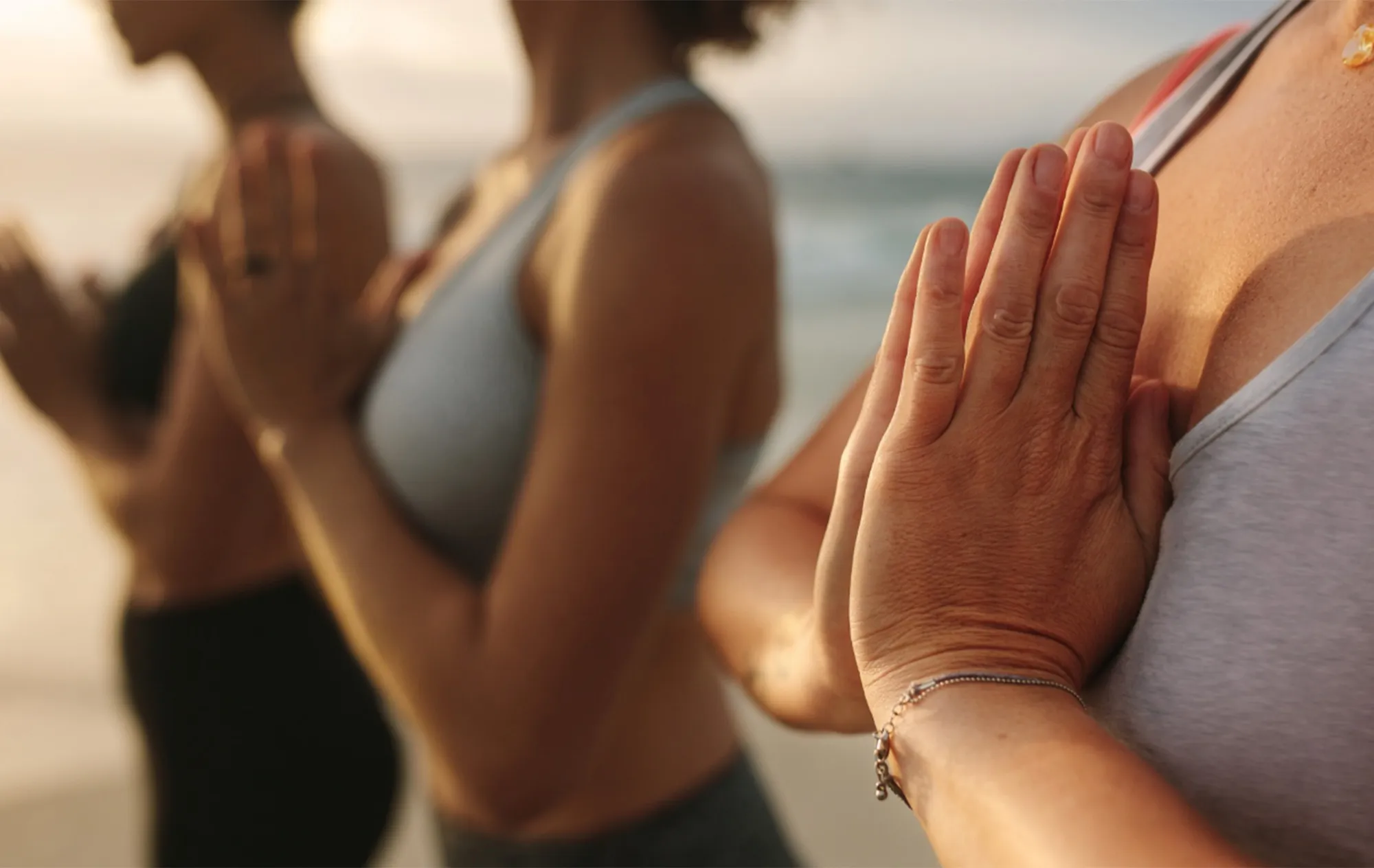 Close-up of 3 women in sports tops standing with their palms pressed together at their chests, the sea blurred behind them
