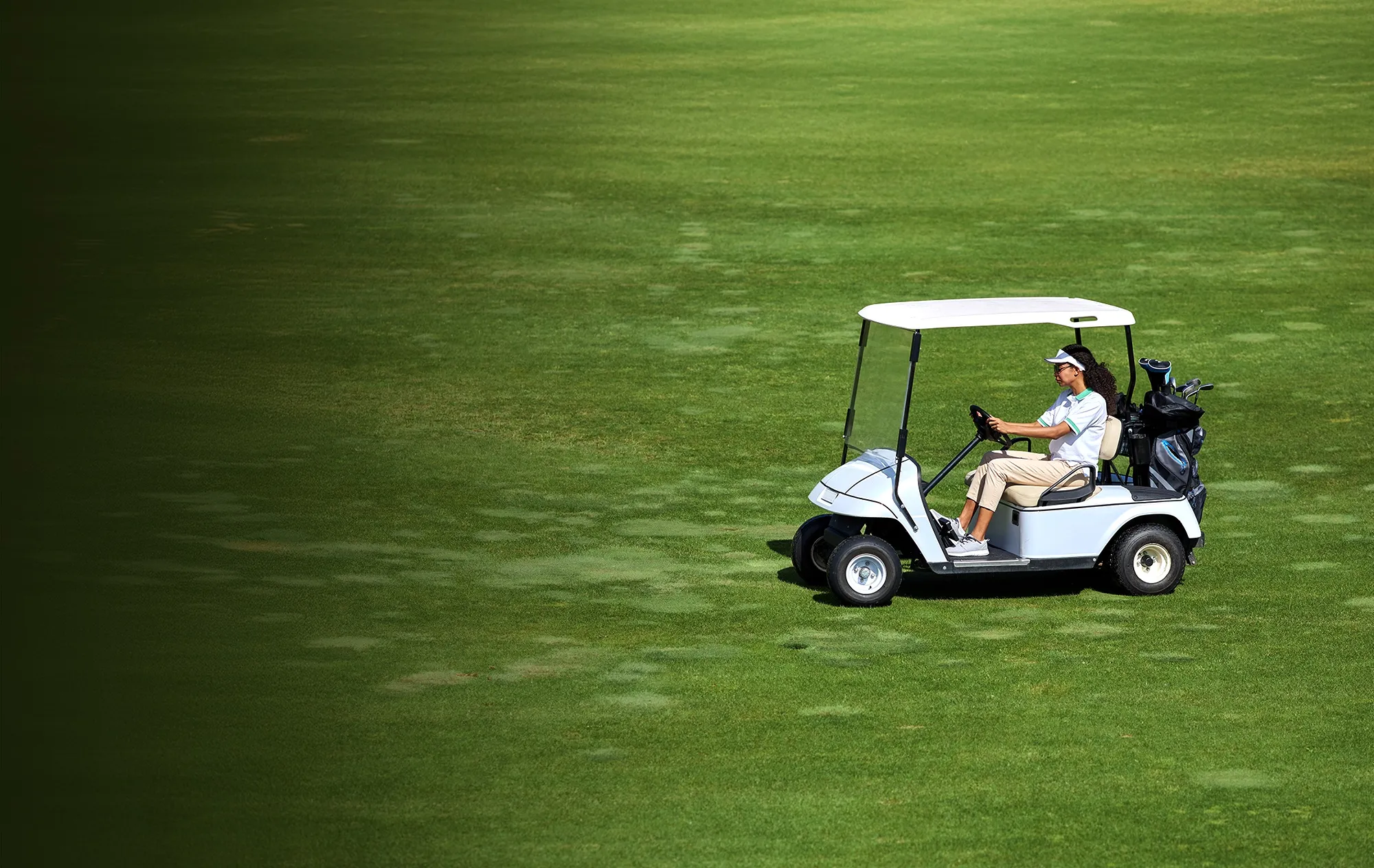 A woman in a white visor driving a white golf cart across a green fairway, a golf bag on the back
