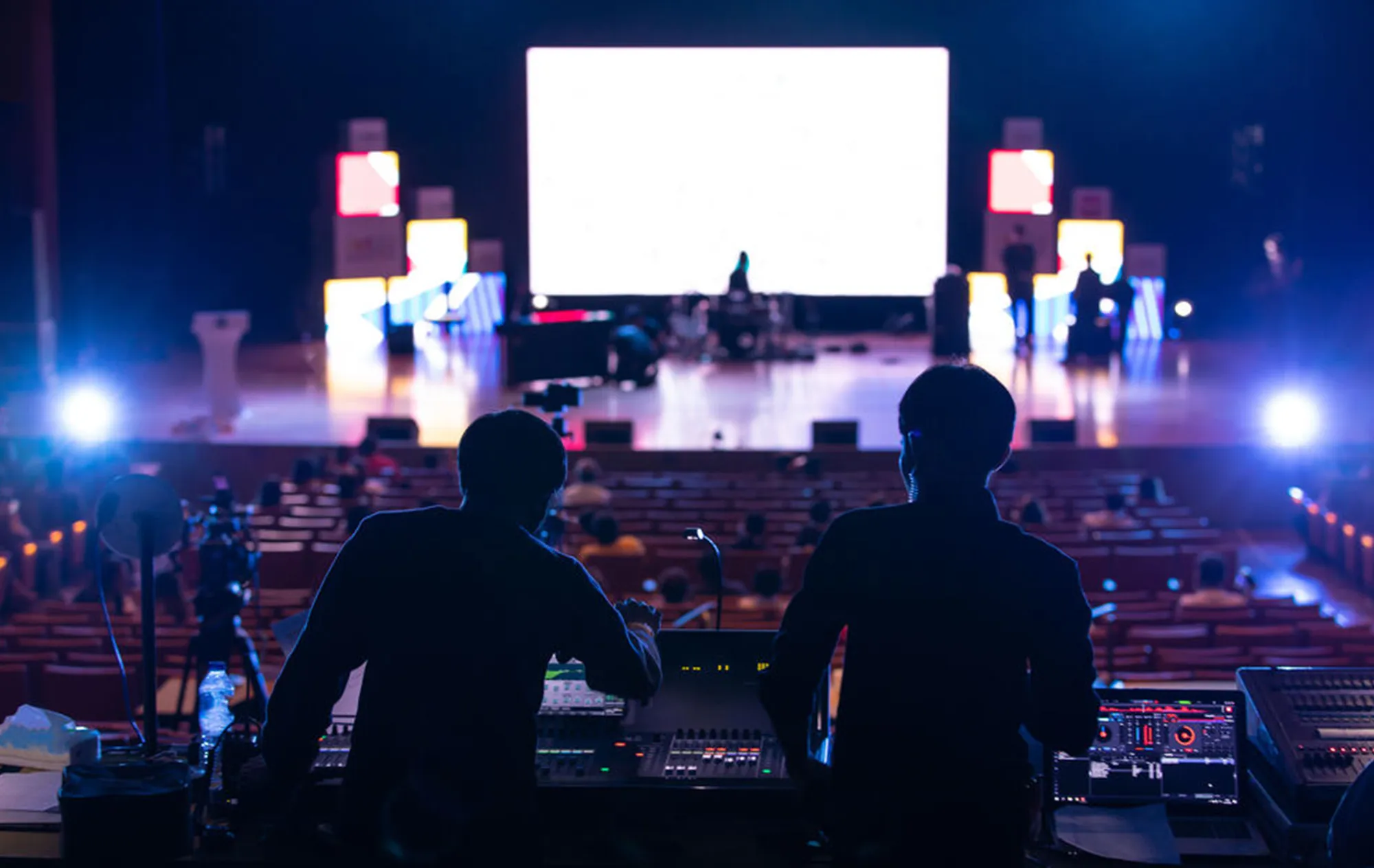Two people in silhouette at a sound and lighting desk, looking across rows of seats to a lit stage with a large white screen