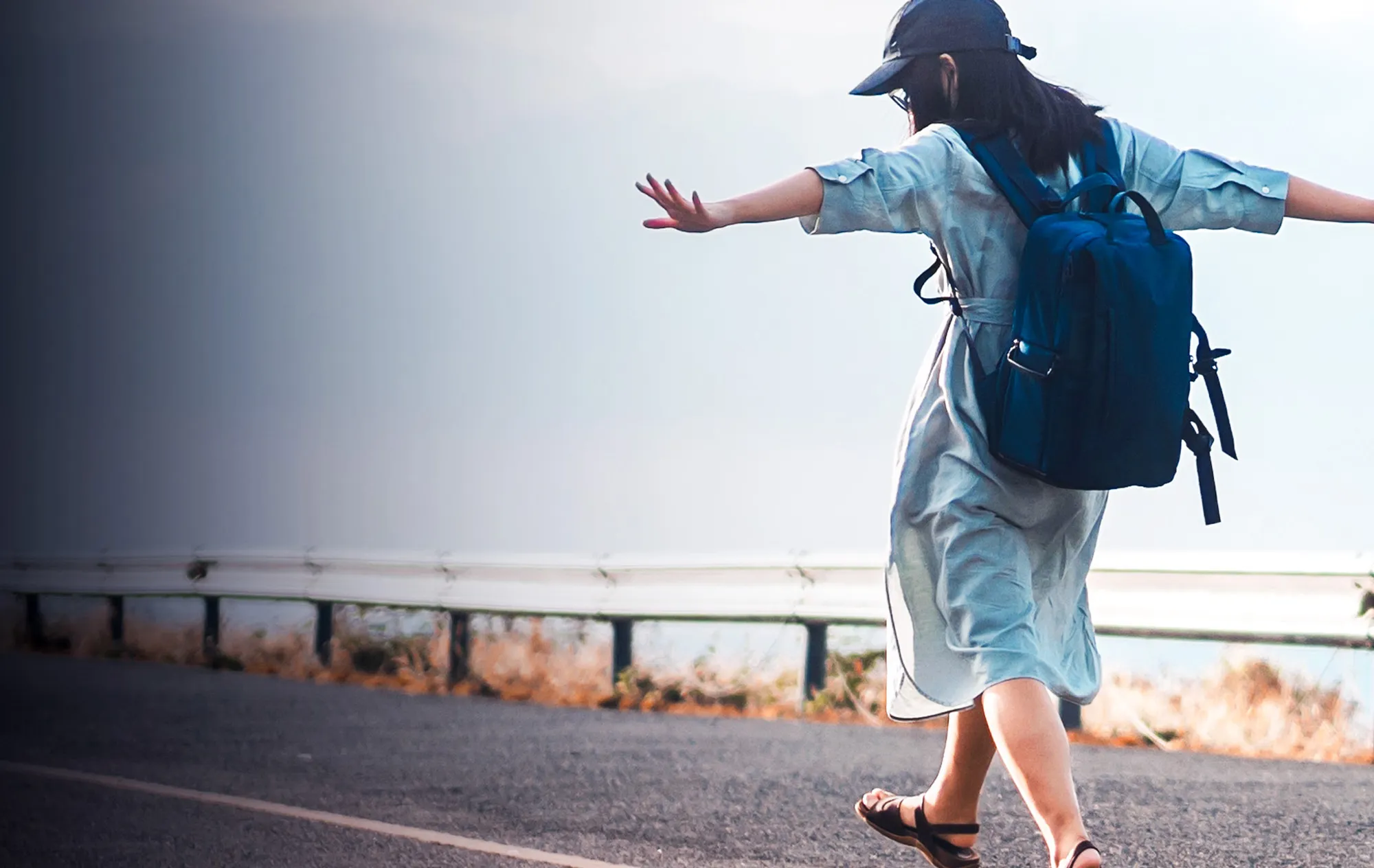 A woman in a pale blue dress, a dark cap and sandals walking along a seaside road with her arms spread wide and a navy backpack on her back