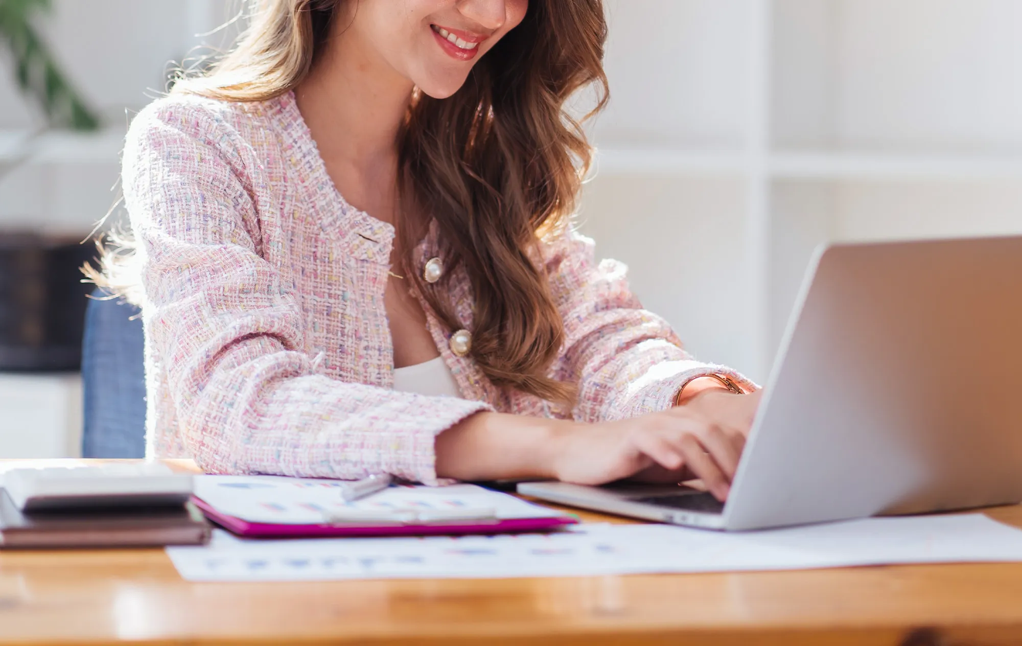 A smiling woman with long wavy hair in a pink tweed jacket typing on a laptop at a wooden desk, printed charts and a clipboard beside her