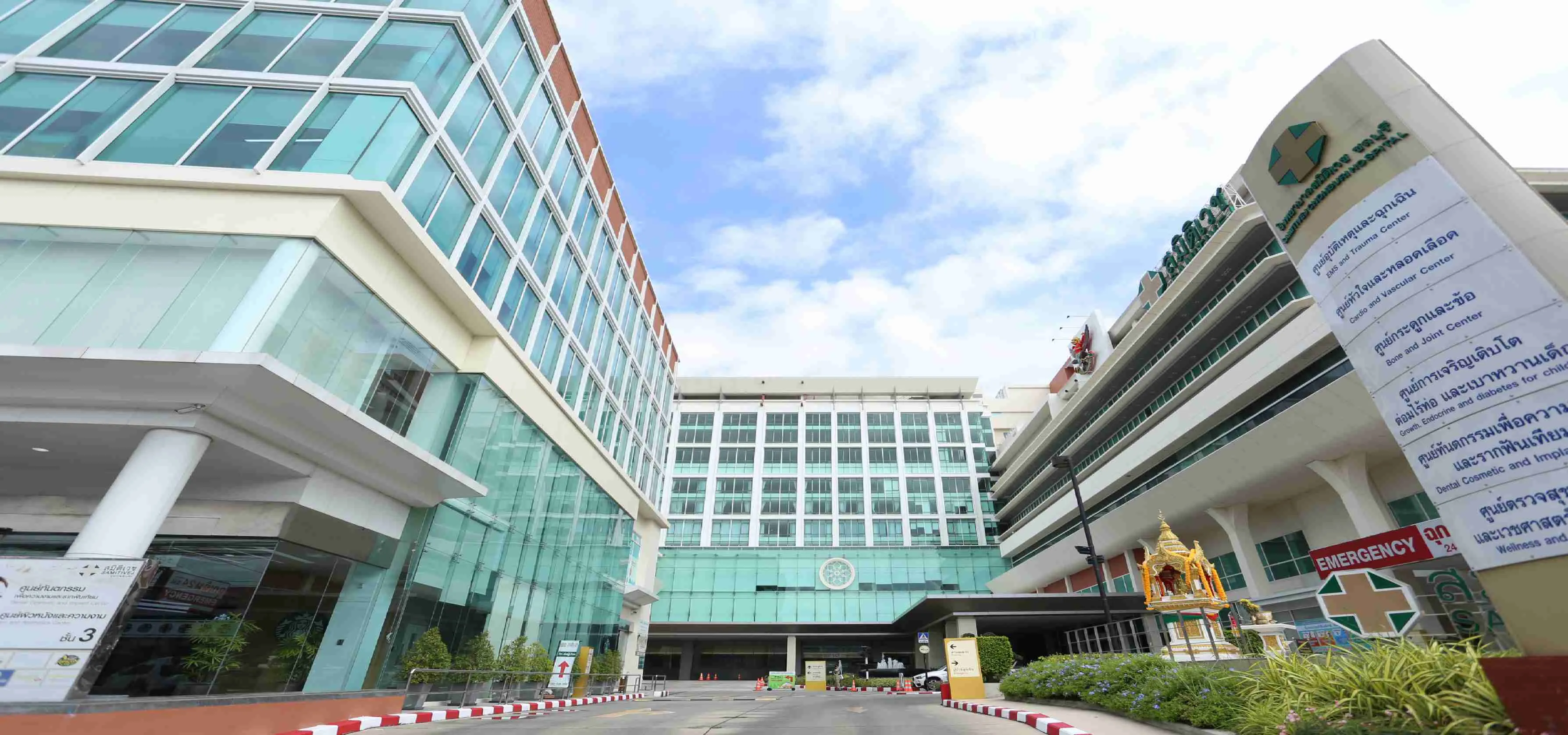The front of Samitivej Chonburi Hospital on a sunny day, with glass-fronted buildings on both sides of the entrance road, the hospital’s green cross and name on the building at the right, a tall sign listing its medical centres and a small golden shrine