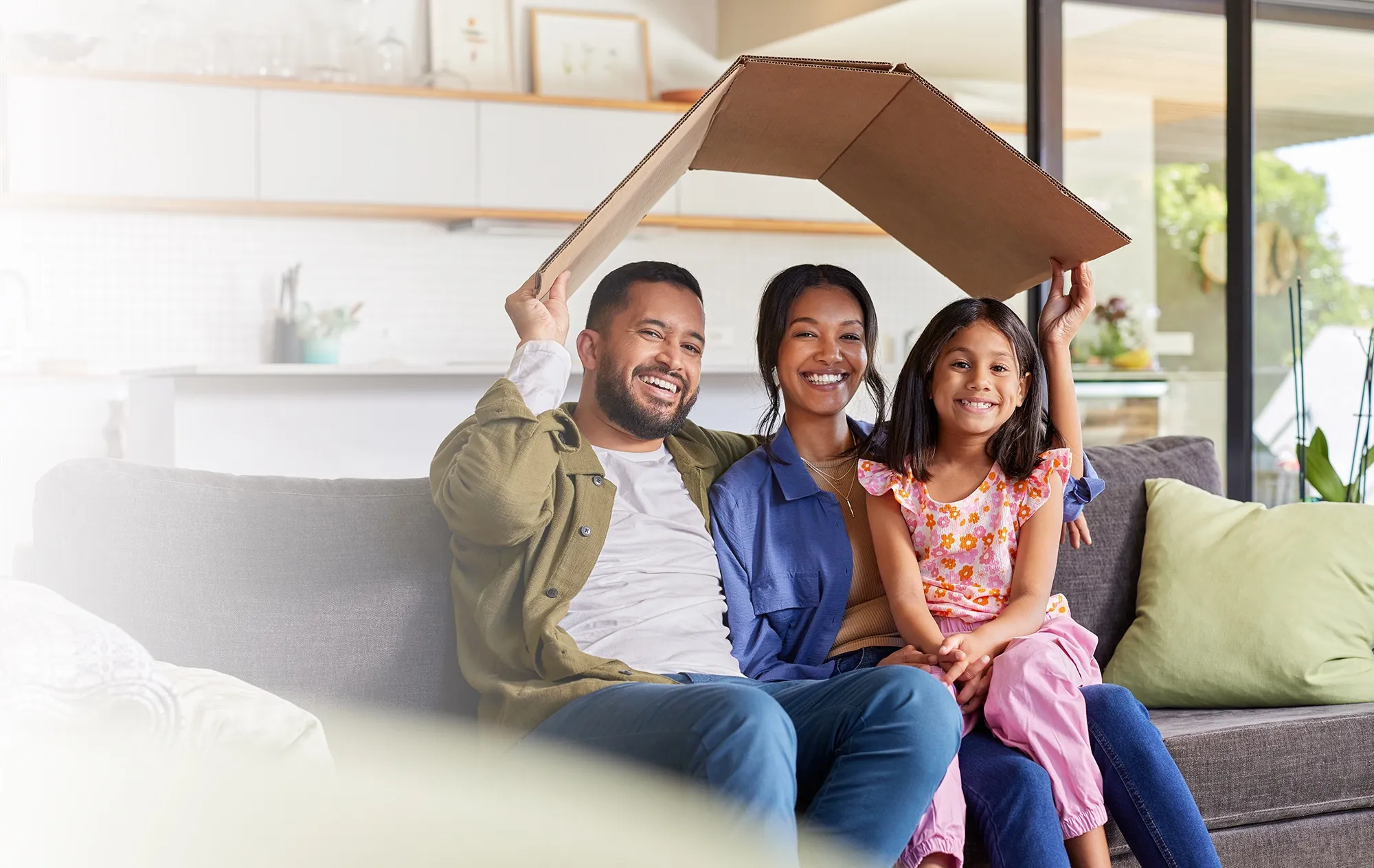 A smiling man, woman and girl on a grey sofa, holding a folded sheet of cardboard above their heads