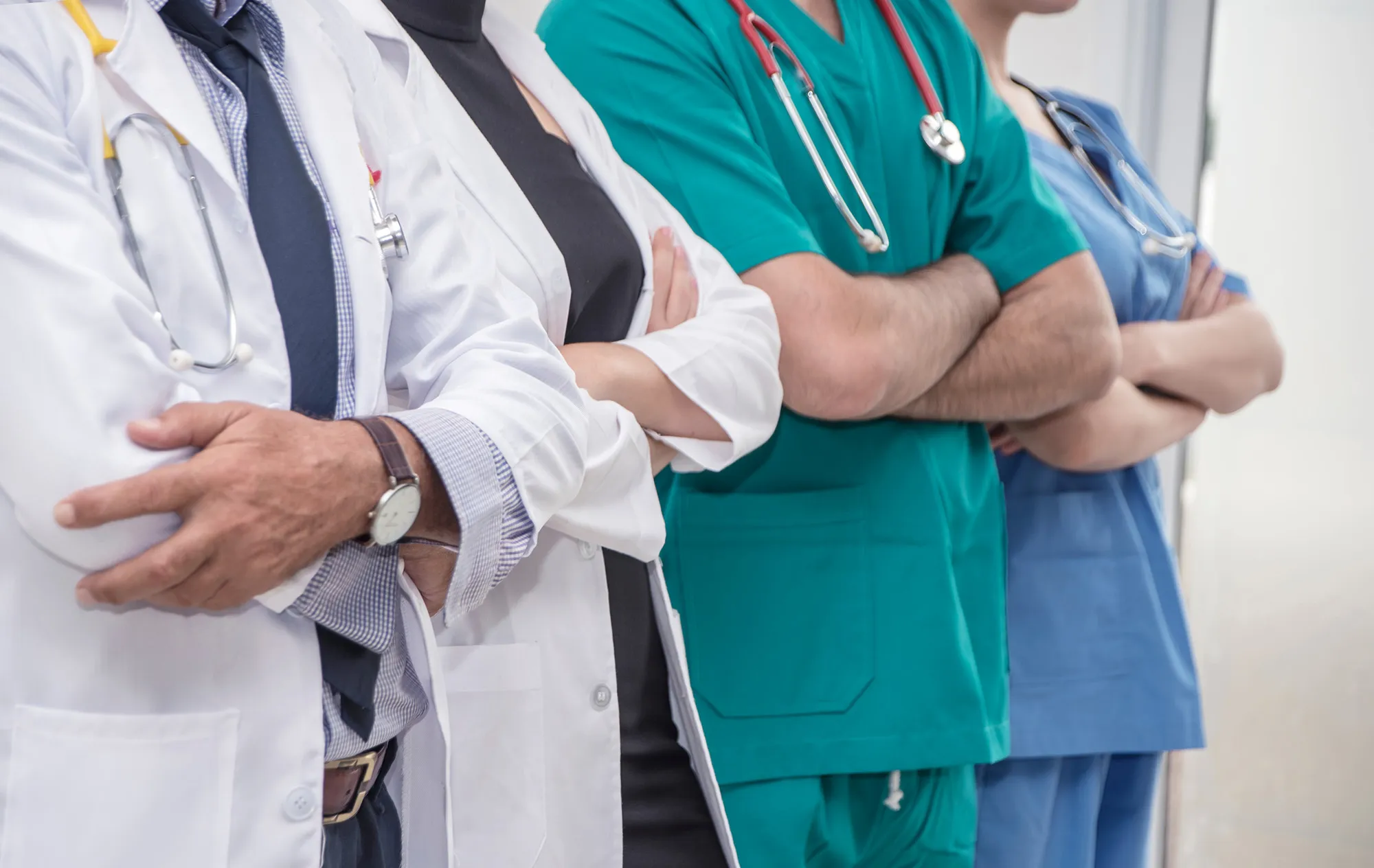 Four medical staff standing in a row with their arms folded, two in white coats and two in green and blue scrubs, three with stethoscopes round their necks