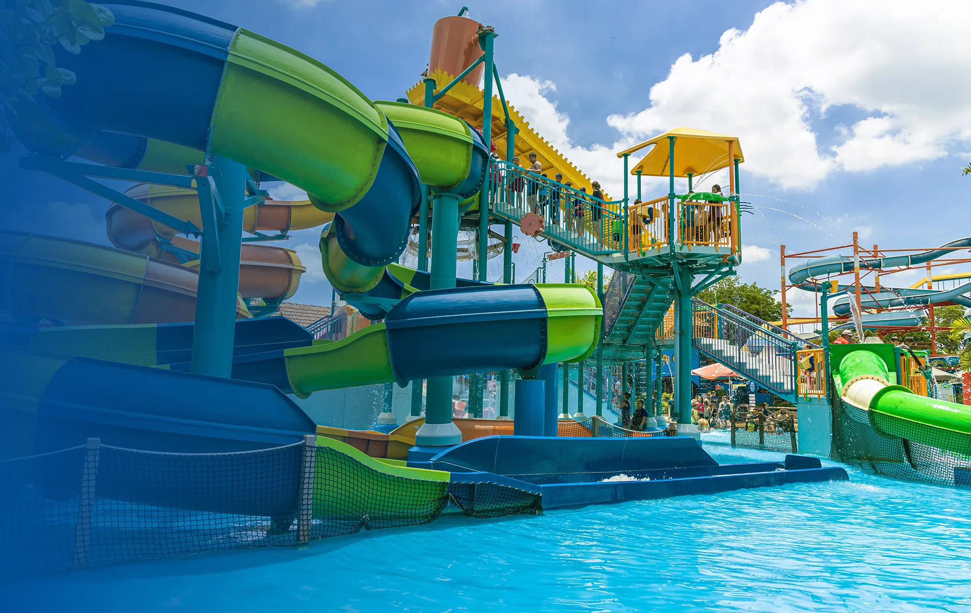 Green, blue and orange water slides twisting above a turquoise splash pool, with people on the stairway up to the slide tower