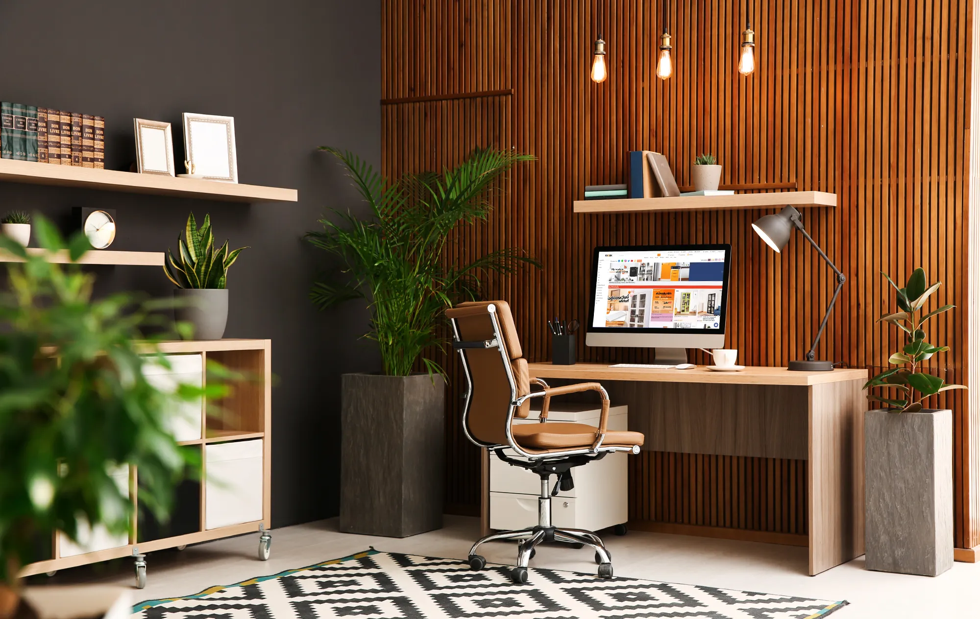 A home office with a slatted wooden wall, a tan leather office chair at a wooden desk and a computer screen showing the KIOSK website, with tall plants on each side