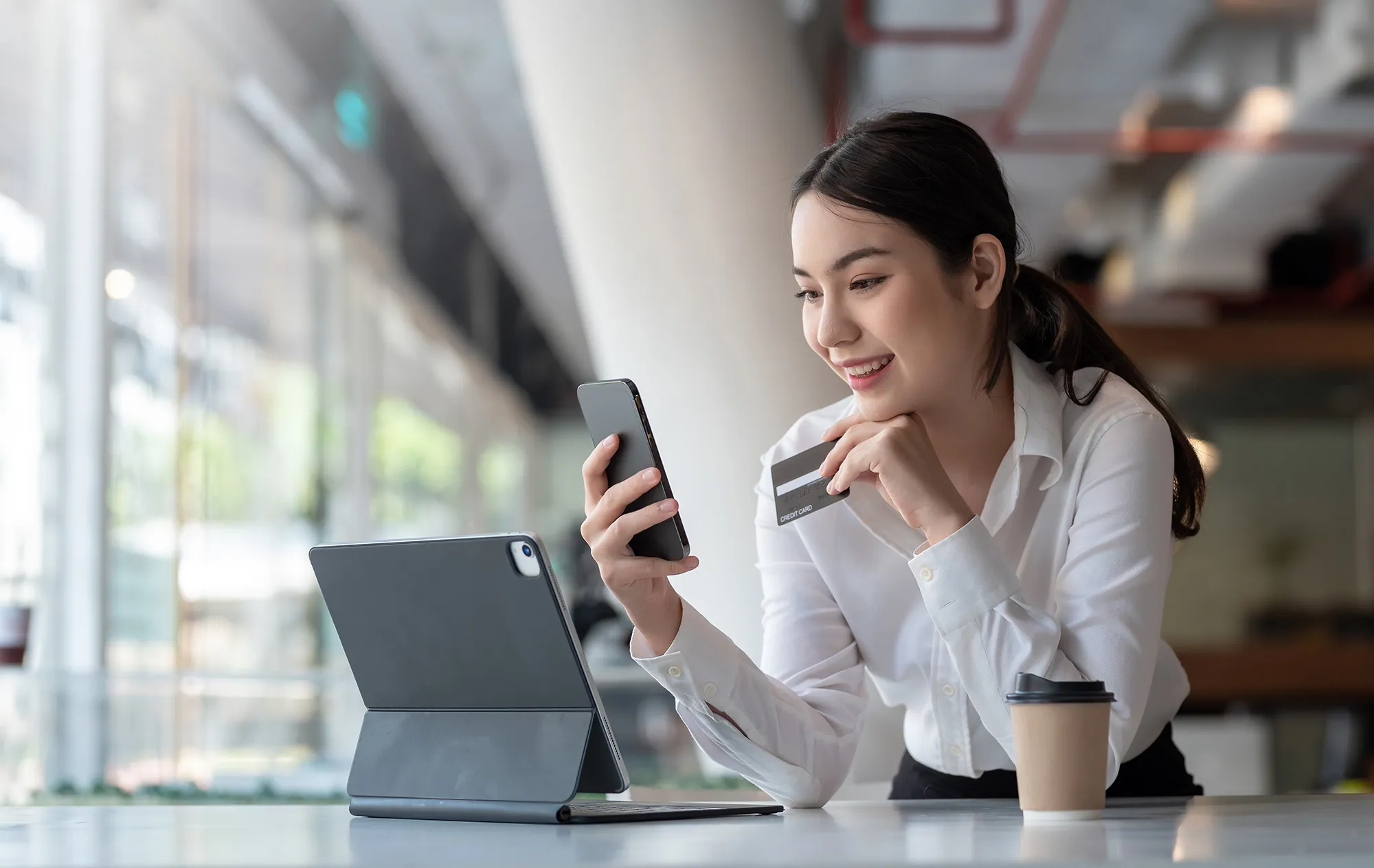A smiling woman in a white shirt holding a phone and a credit card, leaning on a table beside a tablet and a takeaway coffee cup
