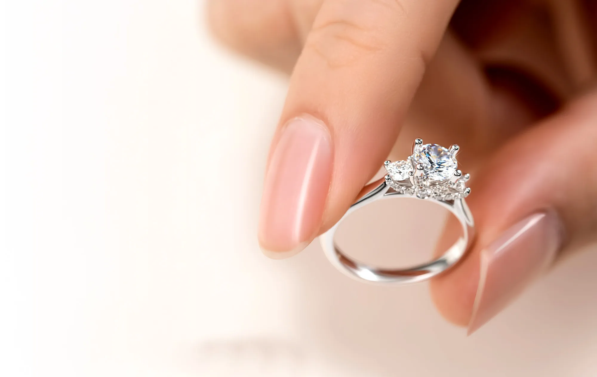 Fingertips holding a ring with a large round stone and smaller stones beside it, against a soft cream background