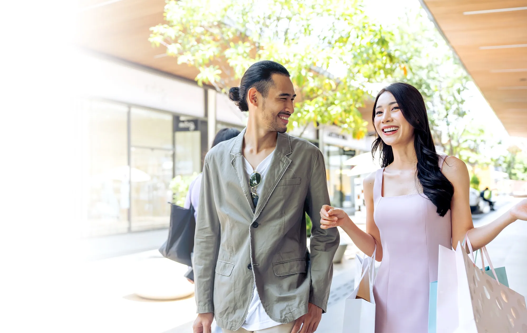 A smiling man in a khaki jacket walking beside a laughing woman in a pale pink dress who carries shopping bags, past a row of shops