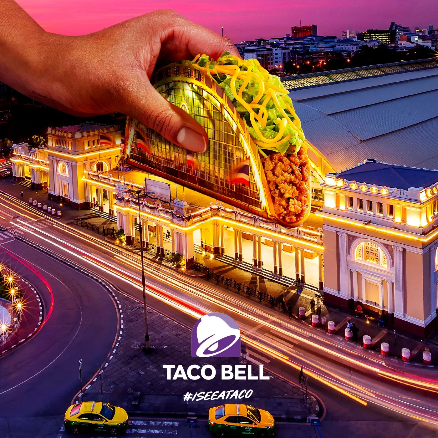 A hand holding a taco whose shell is the arched front of Hua Lamphong railway station at dusk, with taxis below and the Taco Bell logo and #ISEEATACO