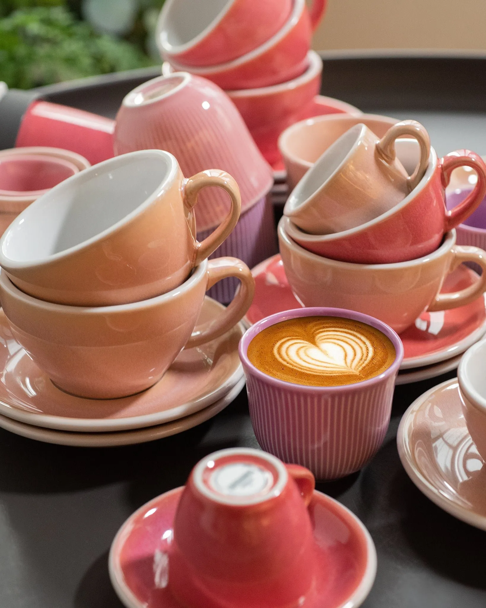 Pink, peach and coral coffee cups stacked on saucers on a dark tray, with a purple ribbed cup at the front holding coffee with a heart in the foam