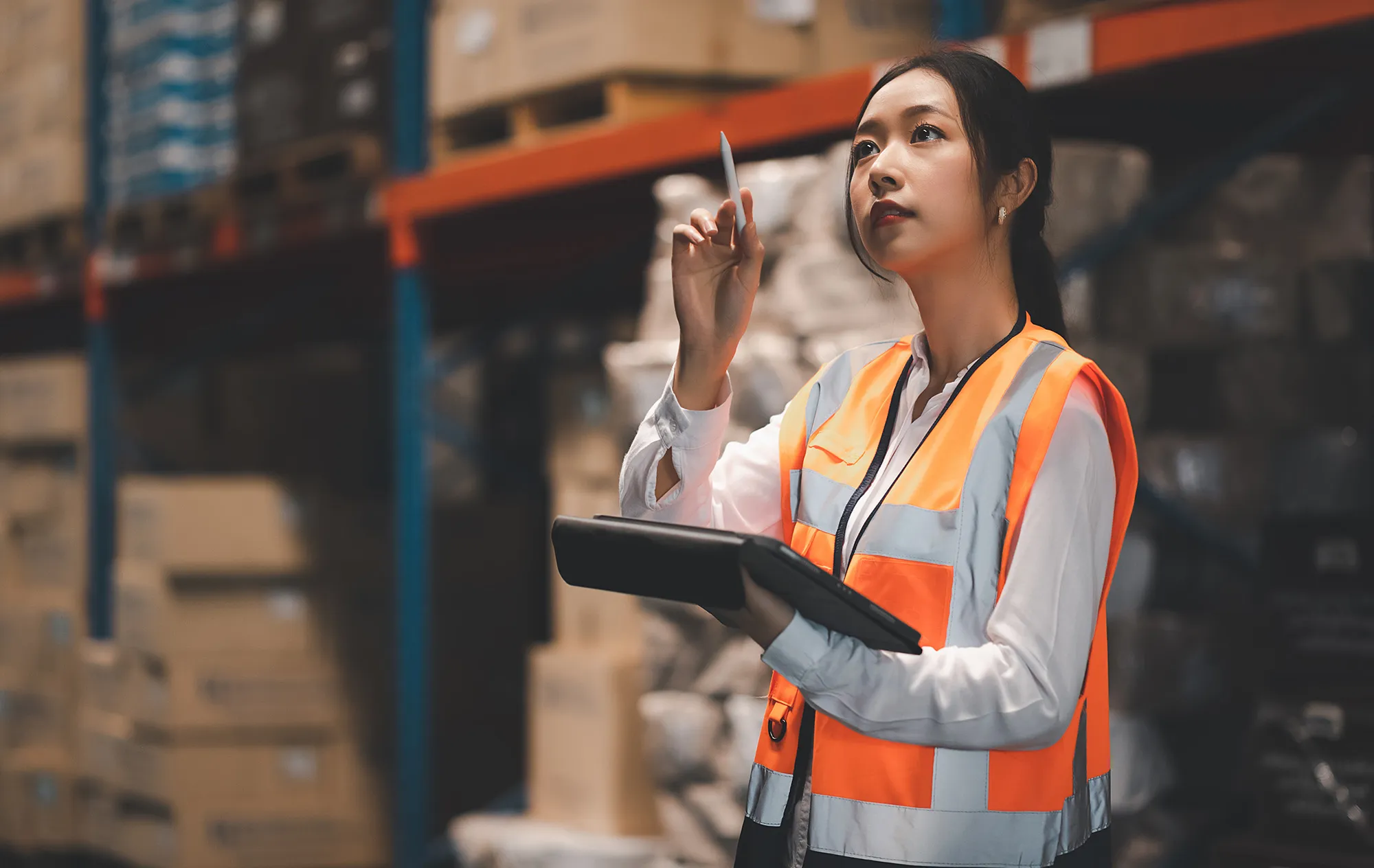 A woman in an orange safety vest holding a tablet and a stylus in a warehouse aisle, looking up at the shelves