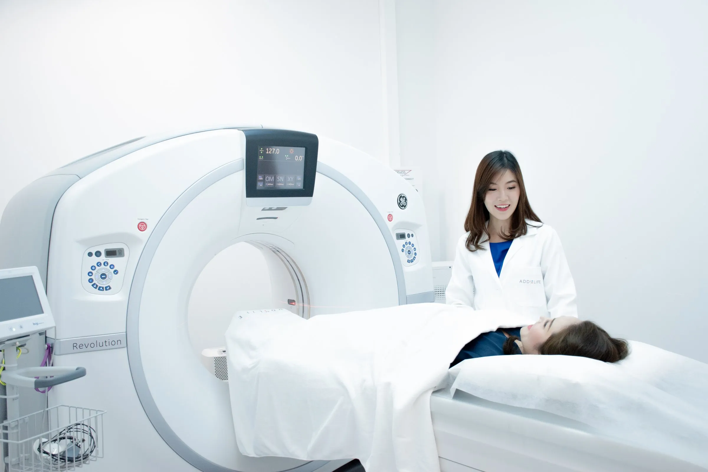 A smiling woman in a white coat with the Addlife logo on the pocket, standing beside a woman lying under a white sheet on the bed of a CT scanner in a bright white room
