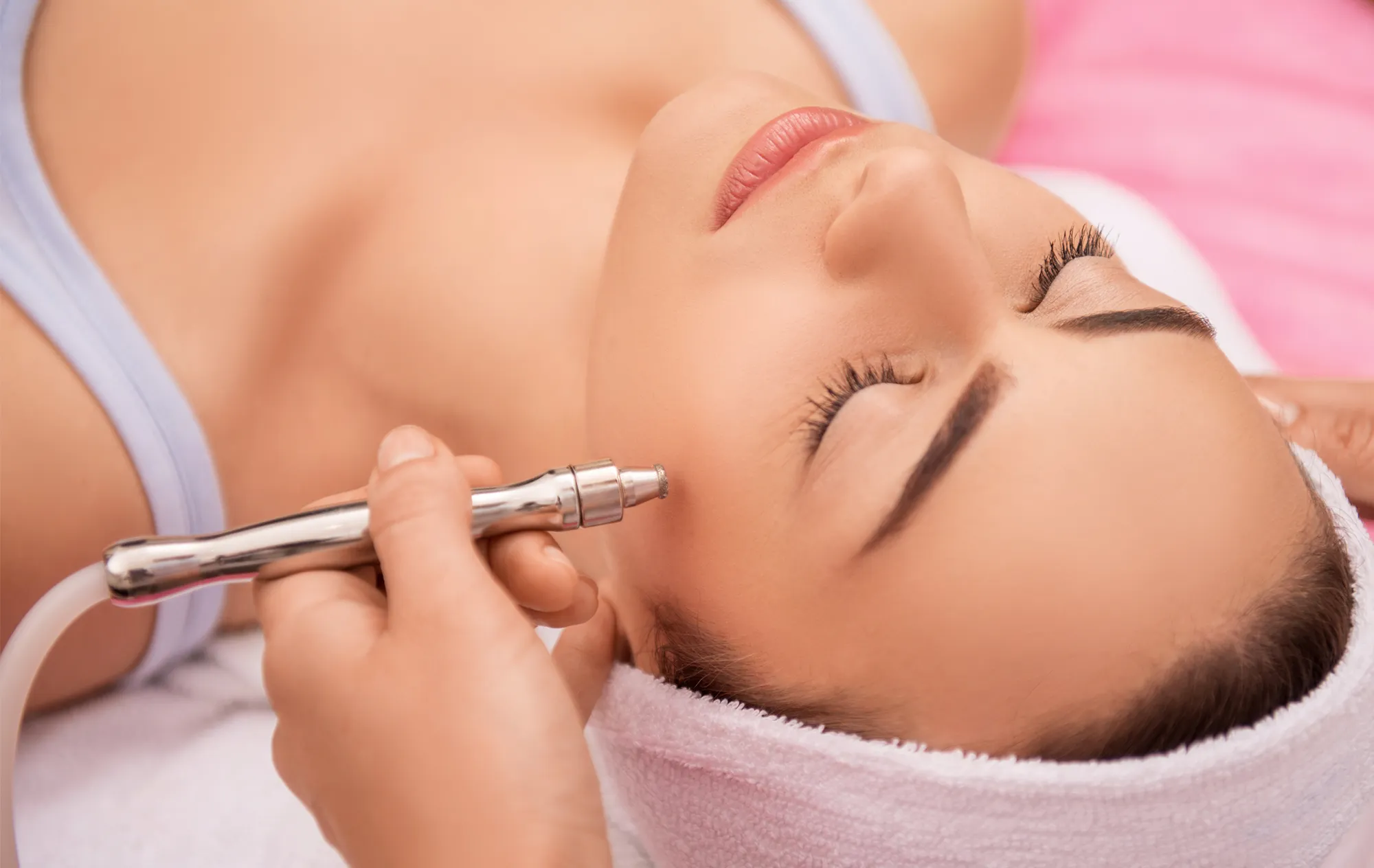 A woman lying with her eyes closed and a towel round her hair while a hand holds a metal handpiece to her cheek