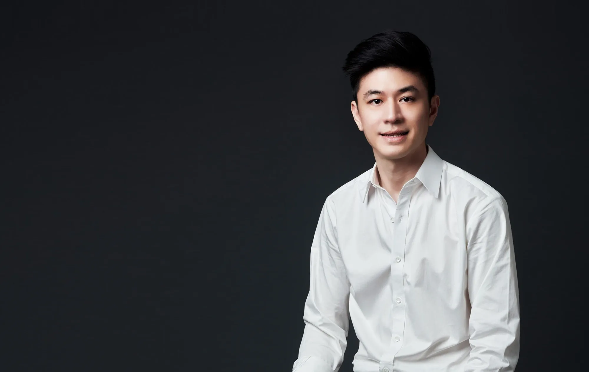 A smiling man with dark swept-back hair in a white shirt against a plain dark grey background