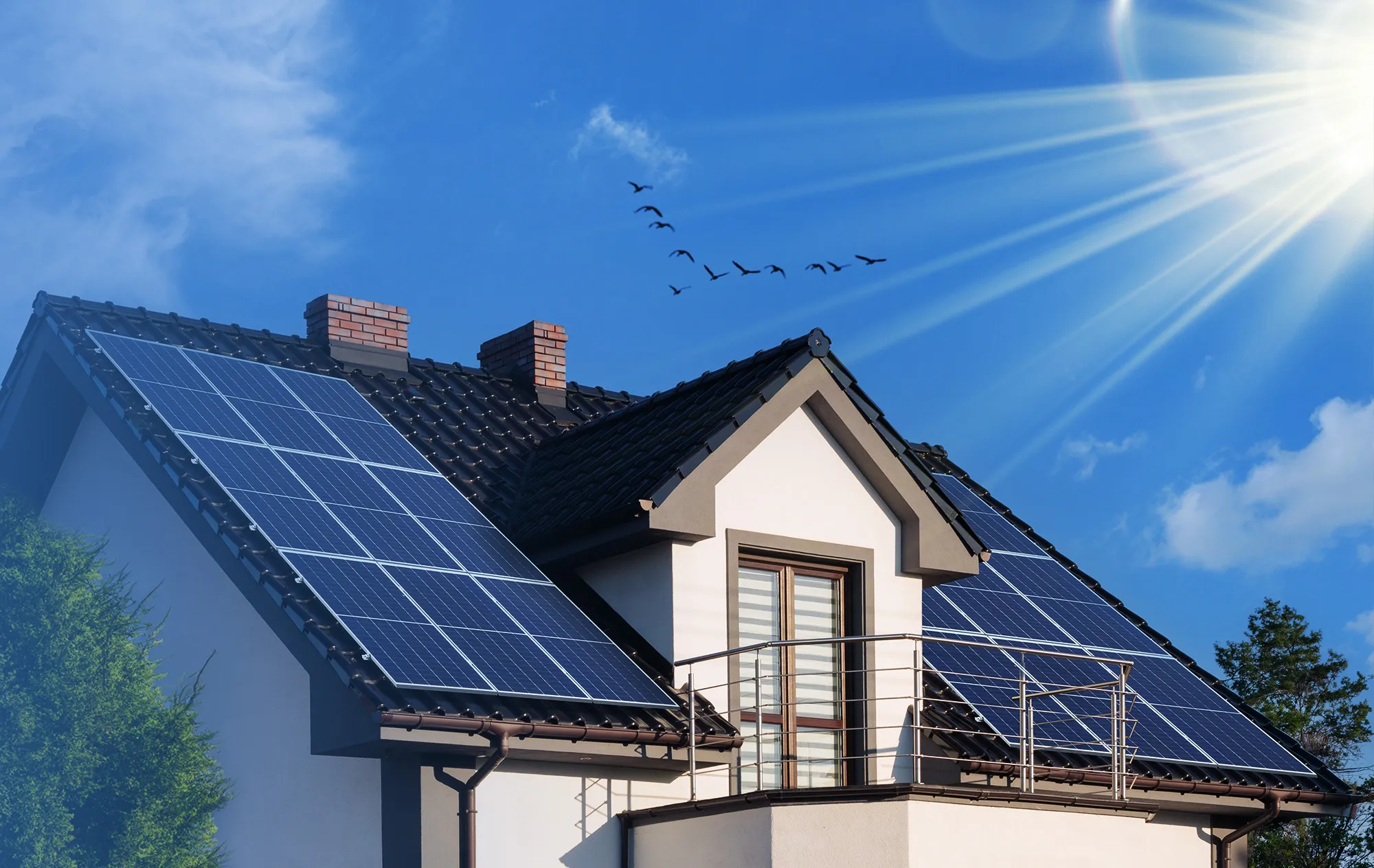 A white house with a dark tiled roof covered in solar panels, under a blue sky with bright sun and a flock of birds