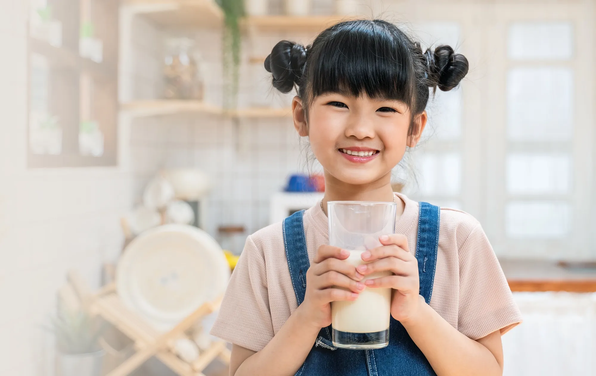 A smiling girl with her hair in two buns holding a glass of milk in both hands in a kitchen