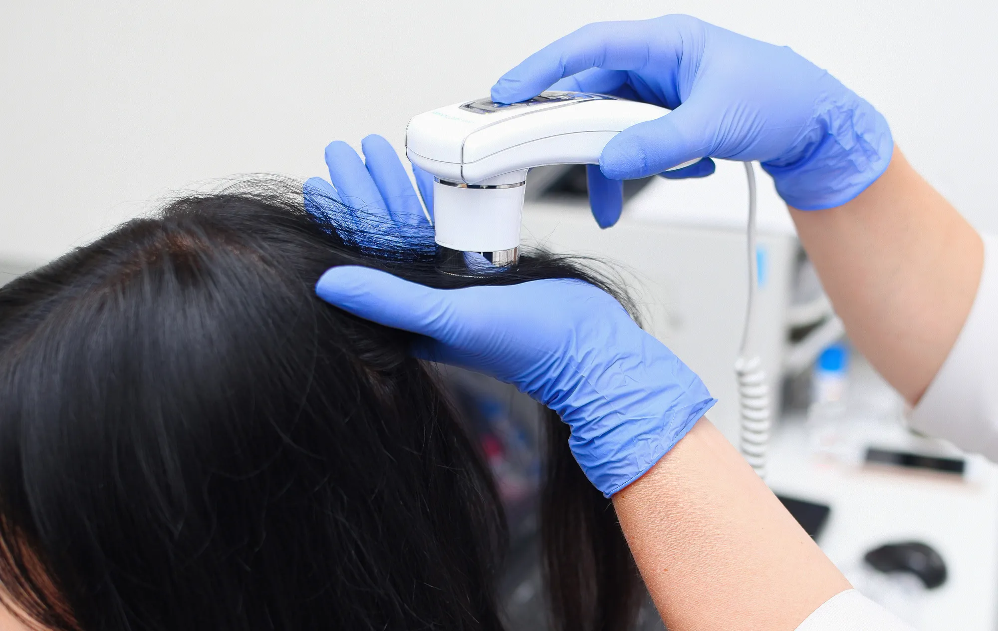 A gloved hand holding a white handheld scanner to the scalp of a woman with long dark hair while the other gloved hand parts her hair