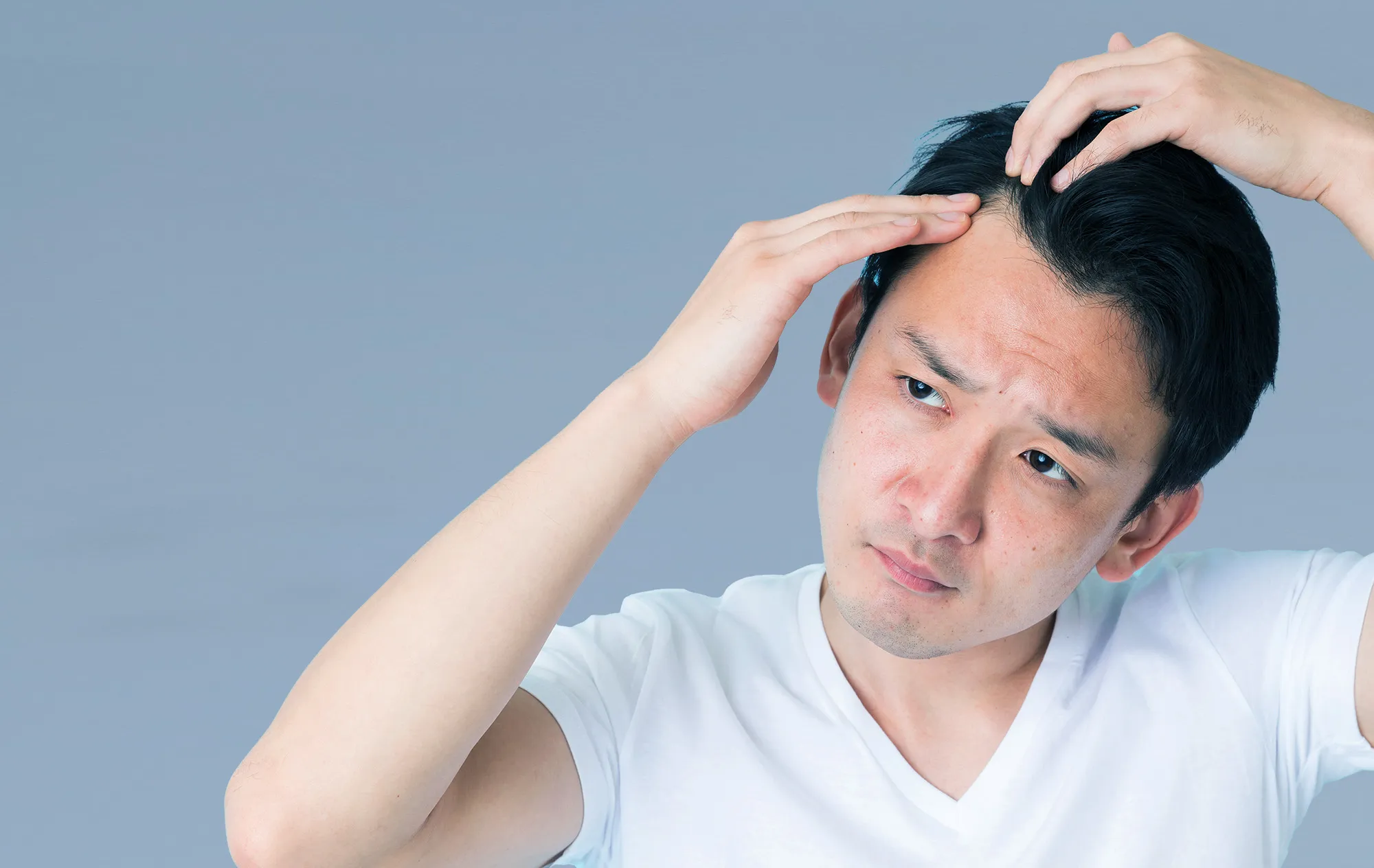 A man in a white T-shirt pushing back his black hair with both hands and frowning, against a grey-blue background