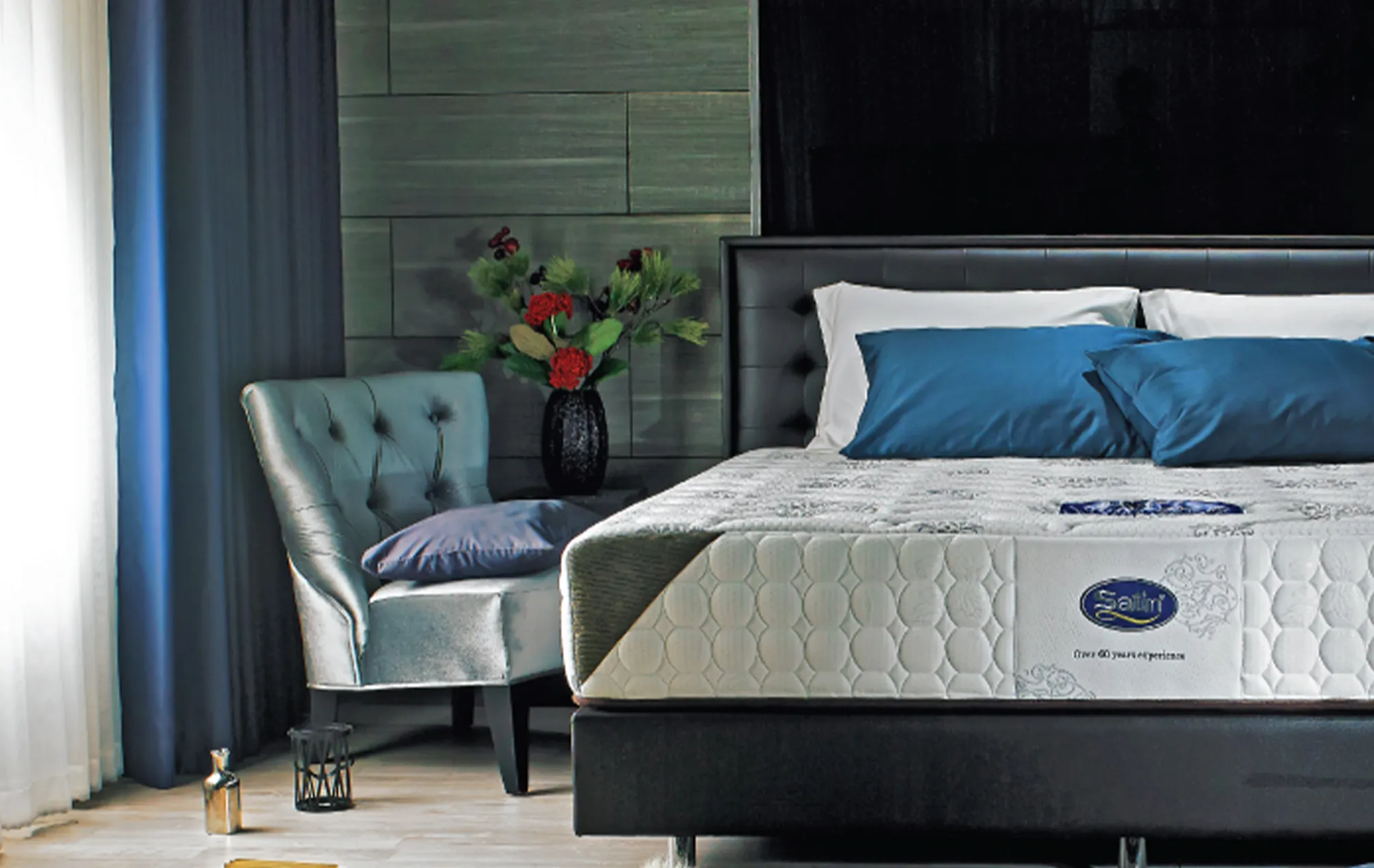 A white quilted Satin mattress on a black leather bed with white and blue pillows, beside a grey armchair and a vase of red flowers