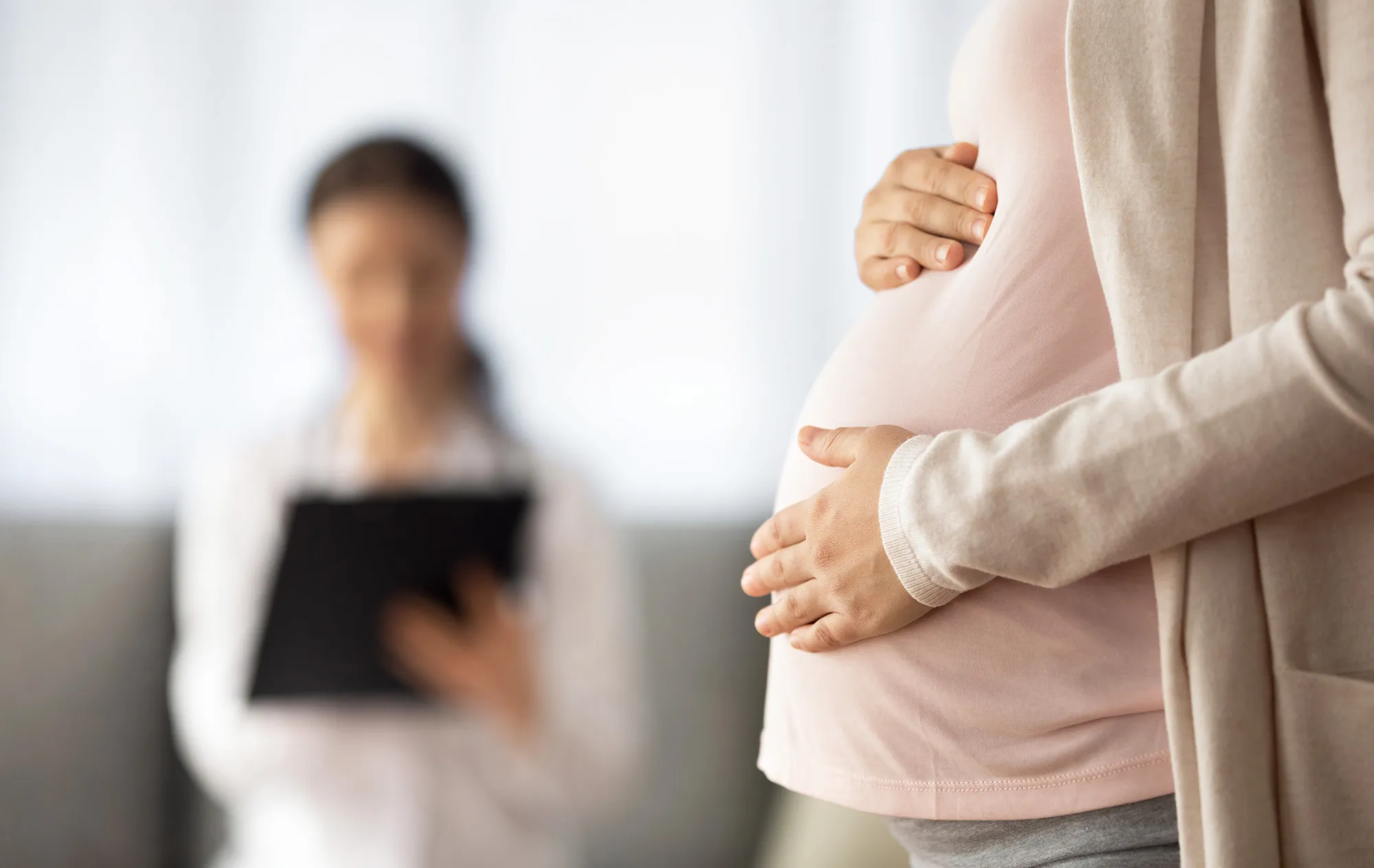 A pregnant woman in a pink top and a beige cardigan holding her belly, a woman in a white coat with a clipboard out of focus behind her