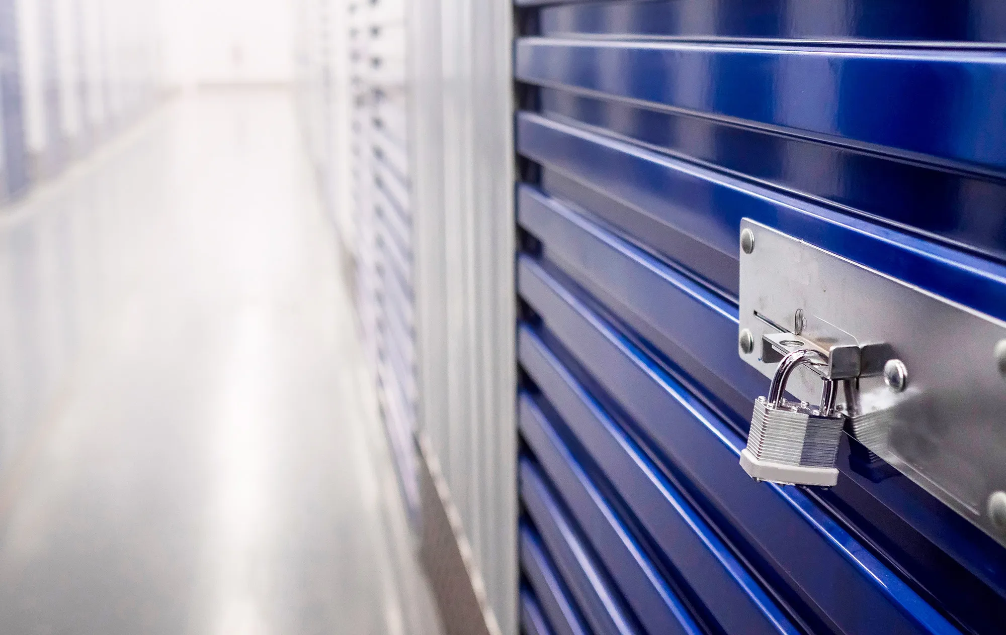 A silver padlock on the latch of a blue roller shutter, with a bright corridor of storage units behind it