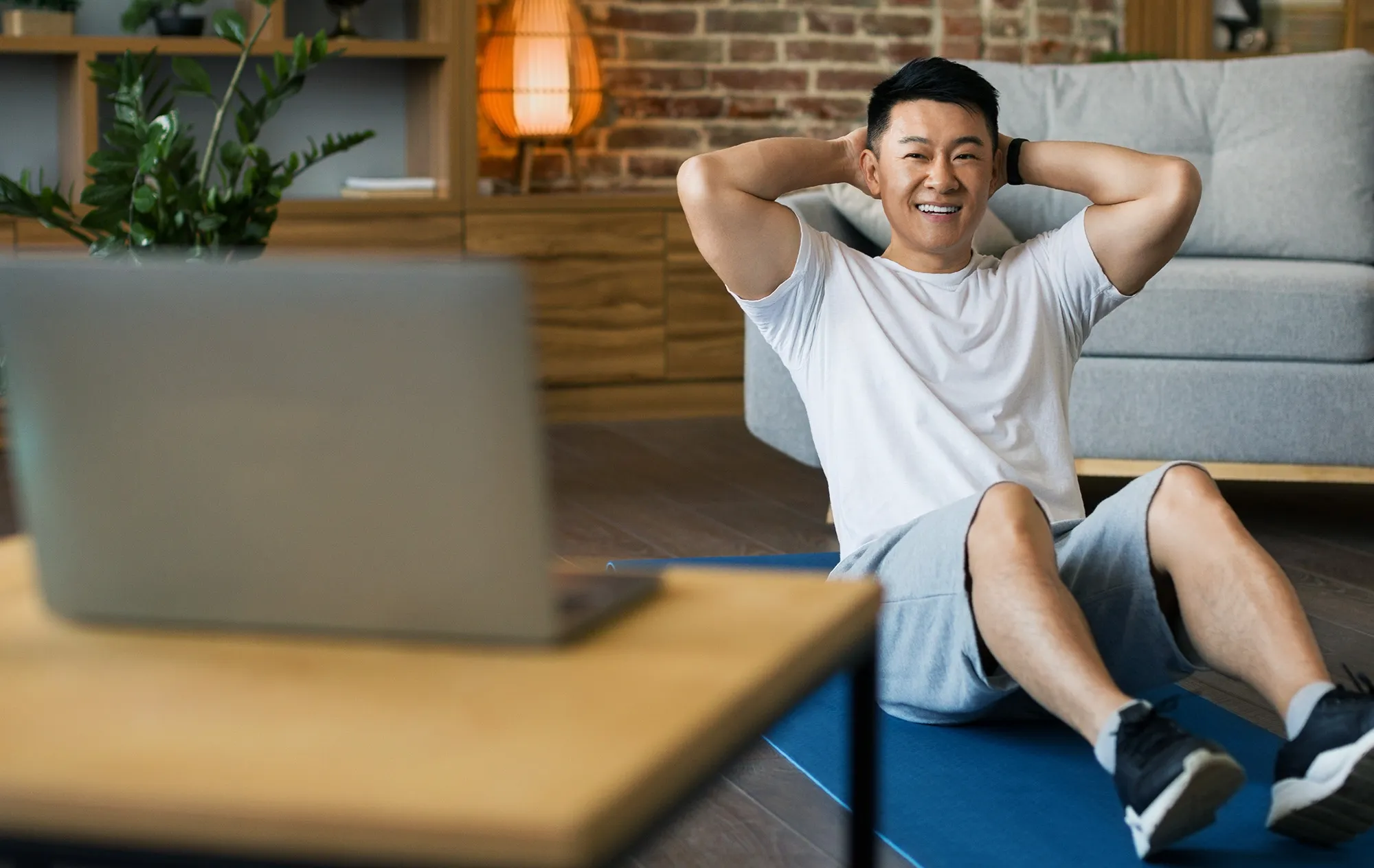 A smiling man in a white T-shirt doing sit-ups on a blue mat at home, a laptop on a table in front of him