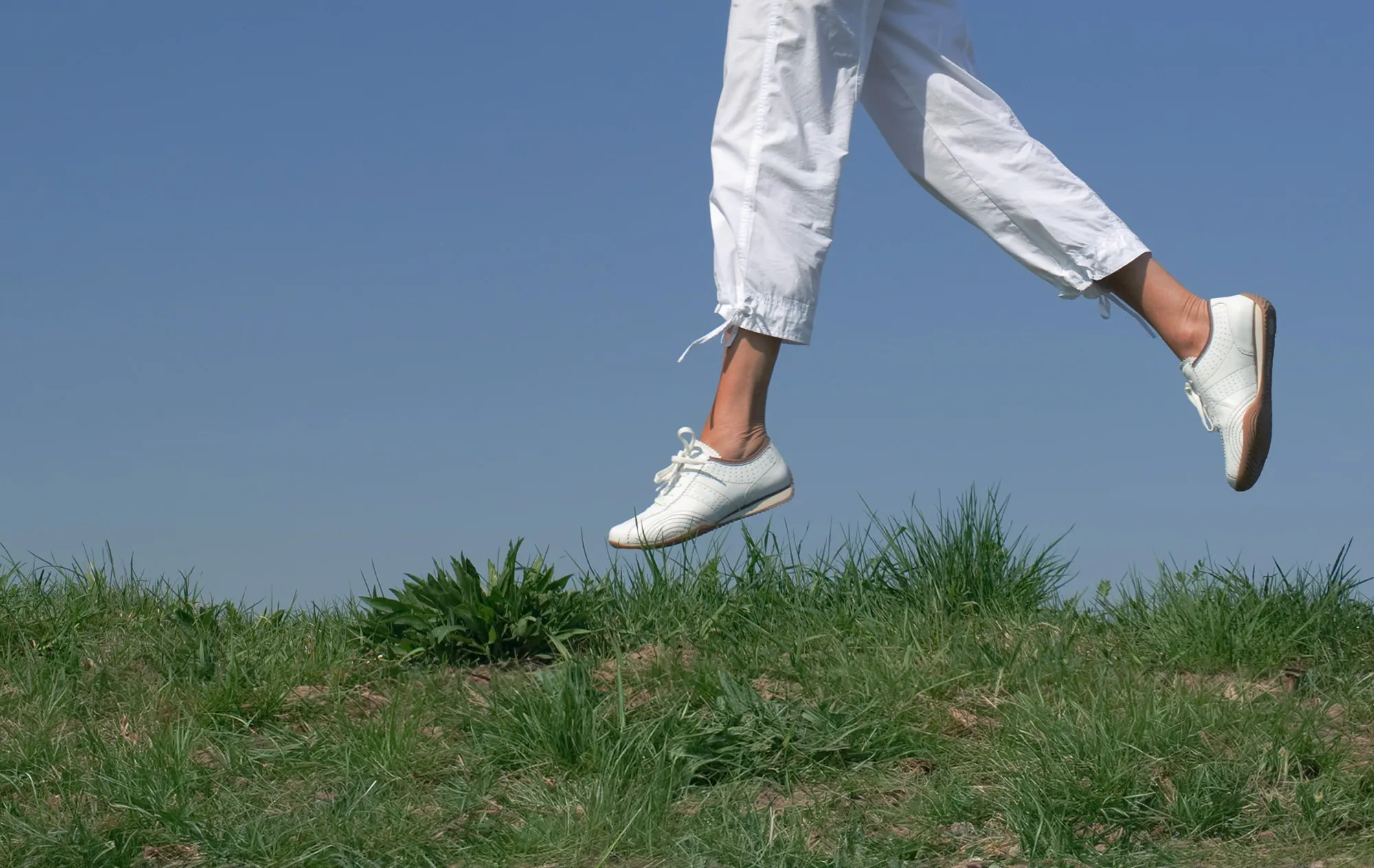 The legs of a person in white cropped trousers and white lace-up shoes, mid-stride above a grassy bank under a blue sky
