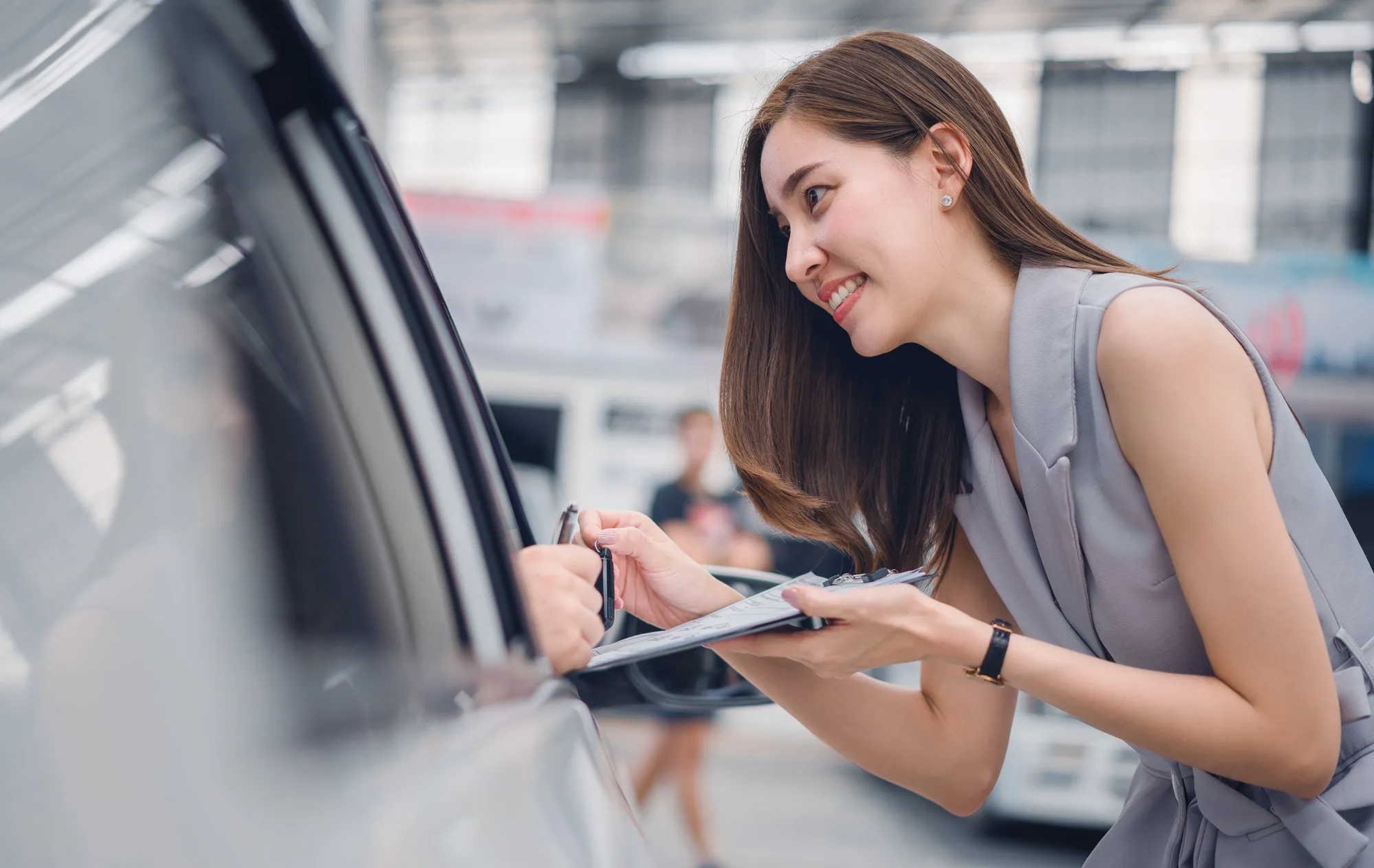 A smiling woman in a grey sleeveless dress leaning towards a car window with a clipboard, passing a car key to the driver