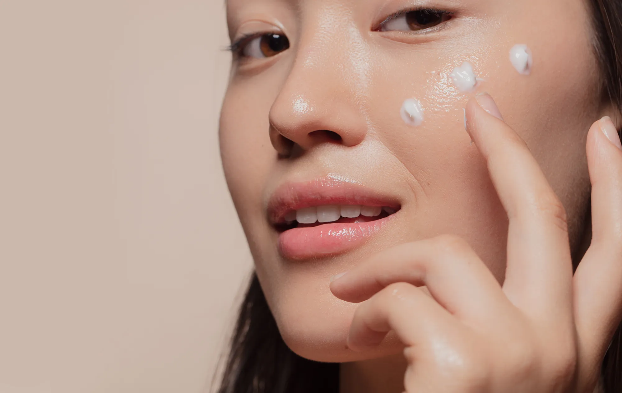 A woman with dark hair smoothing dots of white cream onto her cheek with her fingertips