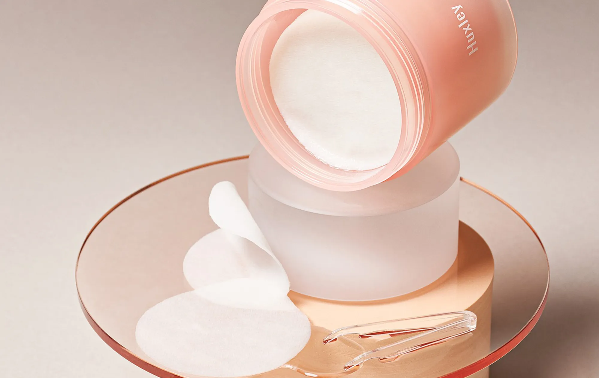 An open pink Huxley jar of cotton pads tilted on a frosted glass block, with loose pads and clear tweezers on a peach acrylic stand