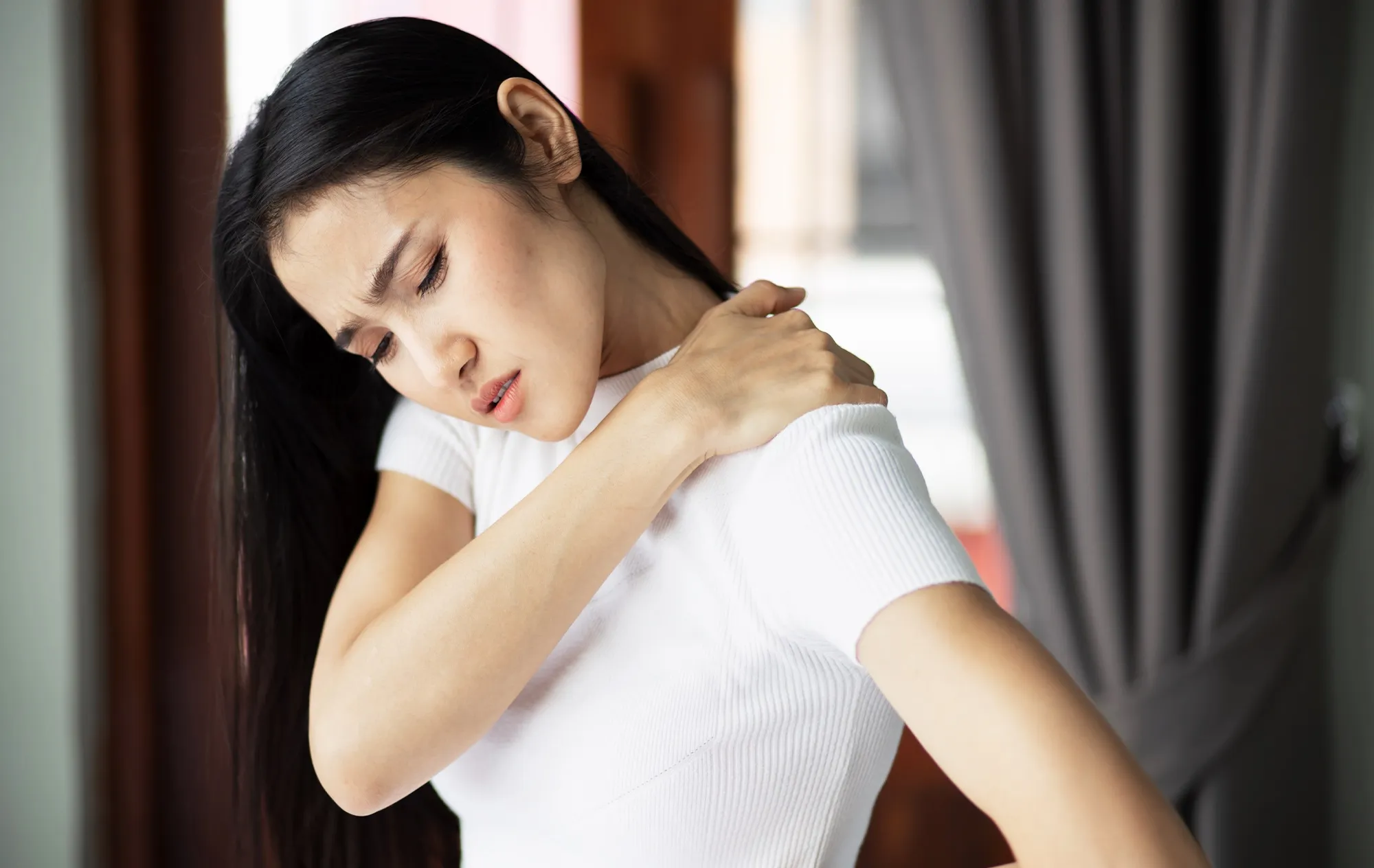 A woman with long black hair in a white T-shirt holding her shoulder, her eyes closed and her brow creased