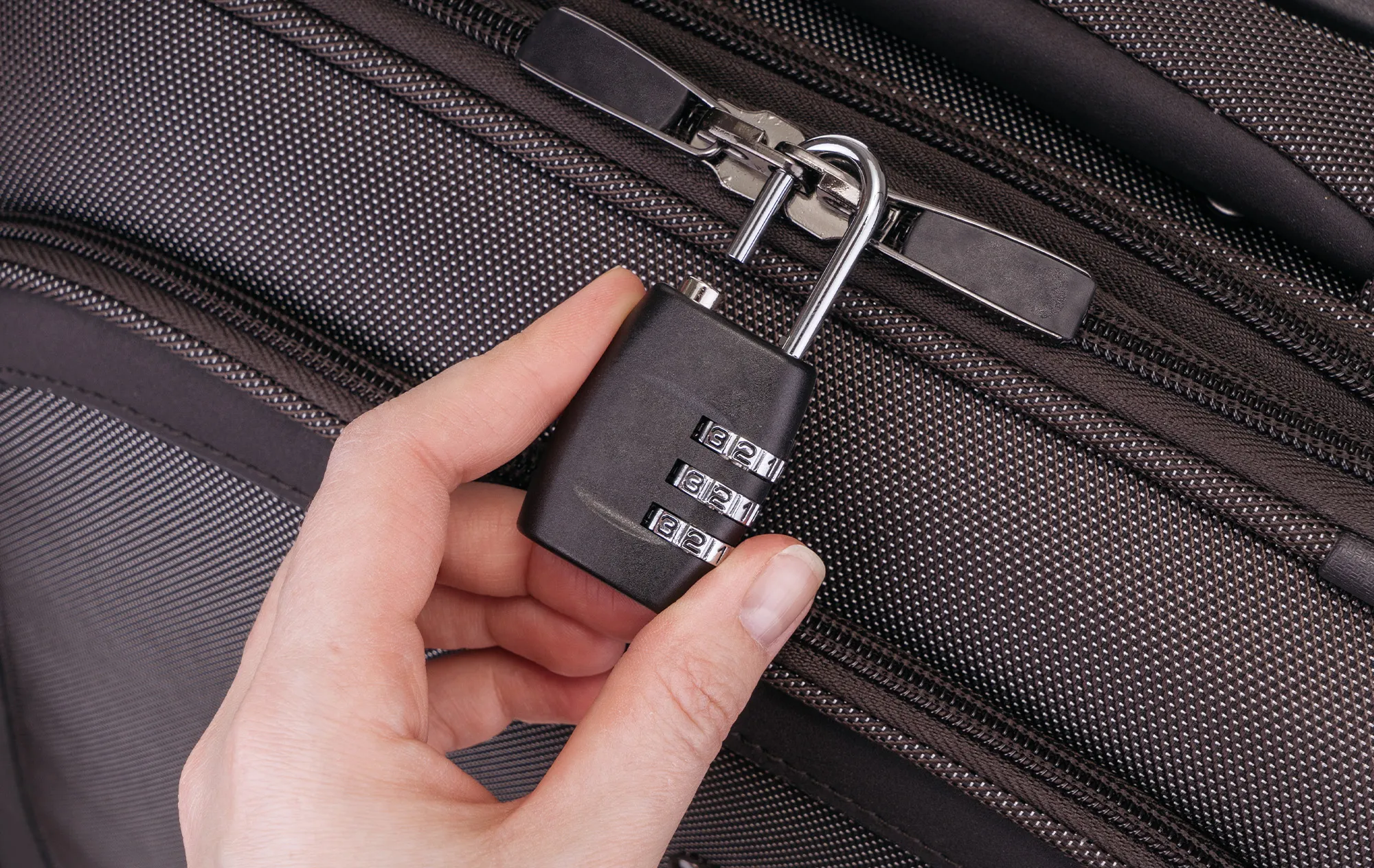 A hand fastening a black combination padlock through the zip pulls of a dark grey bag